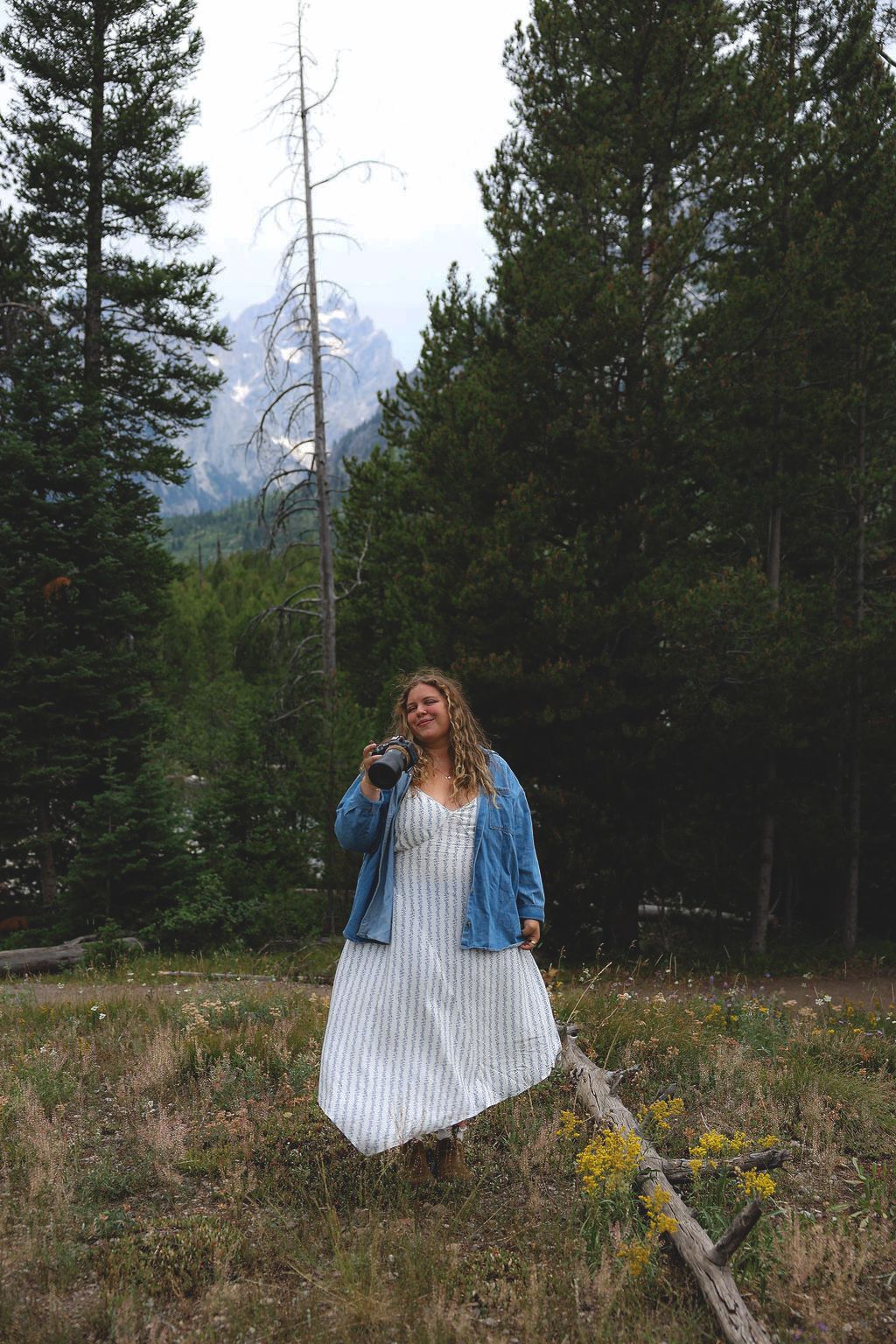 Woman in white dress and denim jacket holding camera in forest meadow with mountain views.