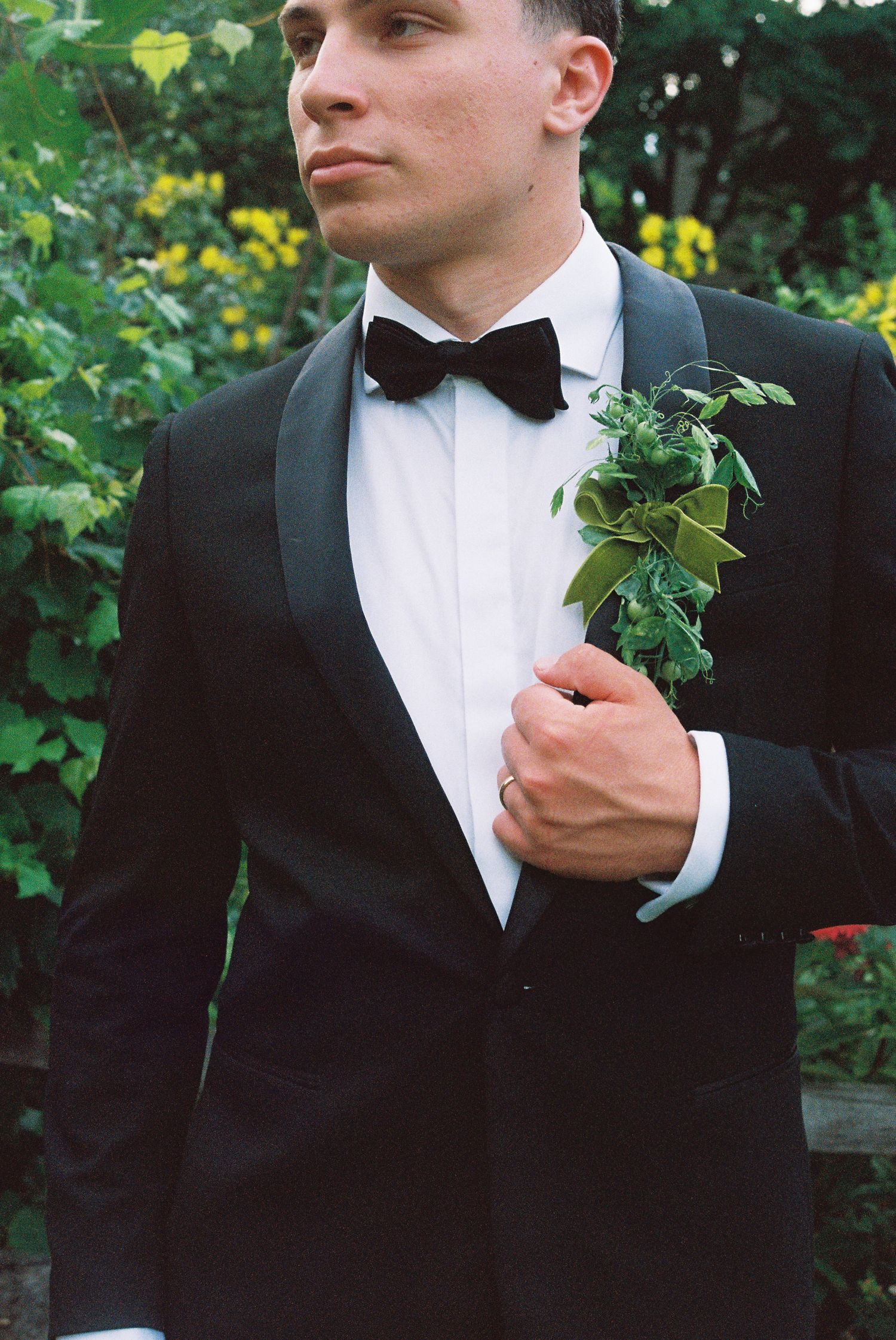 Groom in black tuxedo with velvet bow tie and greenery boutonniere posing outdoors at a wedding.