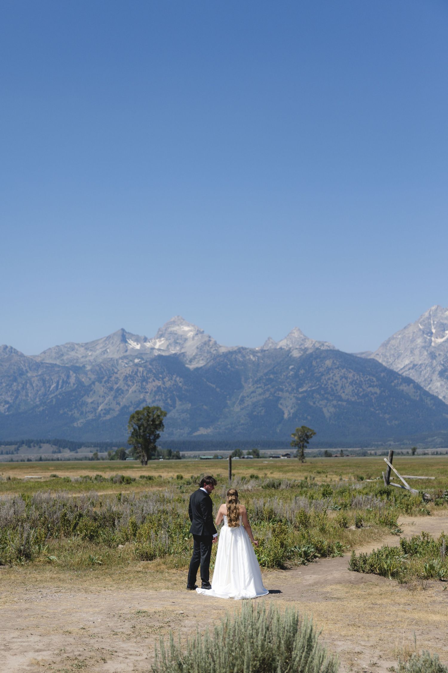 Bride and groom stand together in Grand Teton National Park with majestic mountain range backdrop on clear sunny day.