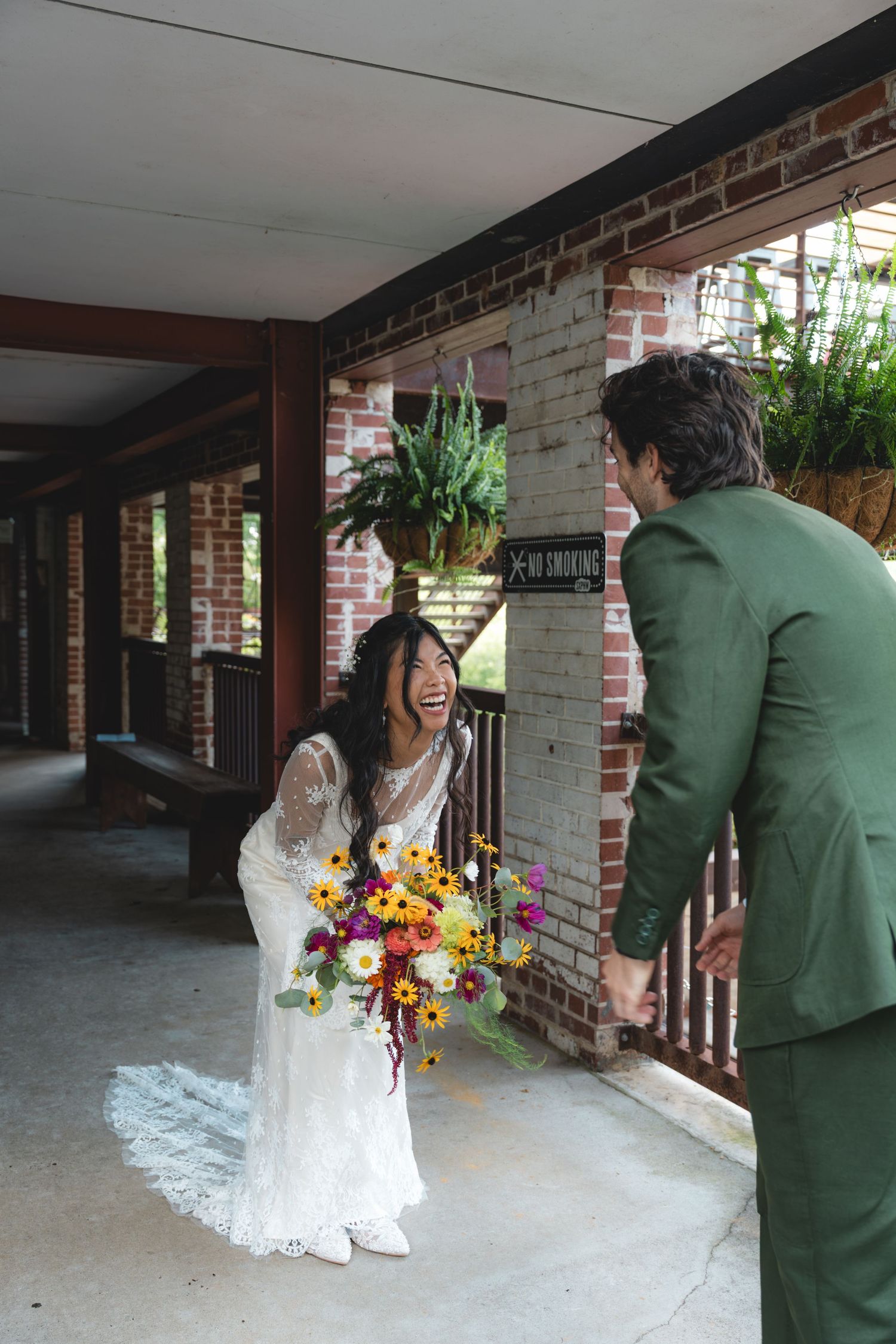 Bride in white lace gown holding colorful bouquet laughs with groom in green suit during outdoor first look.