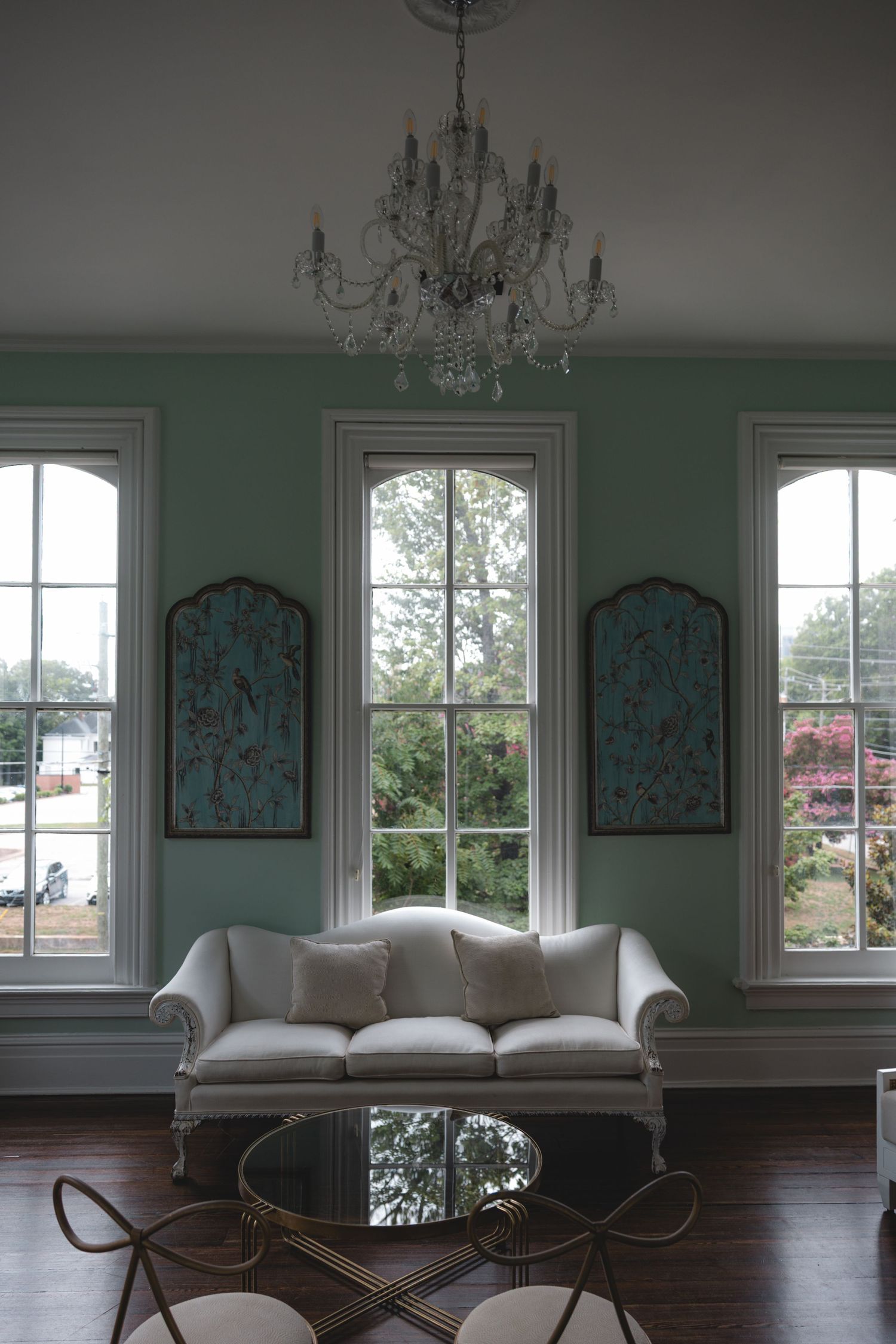 Elegant living room with mint green walls, white camelback sofa, crystal chandelier, and arched windows overlooking a garden.