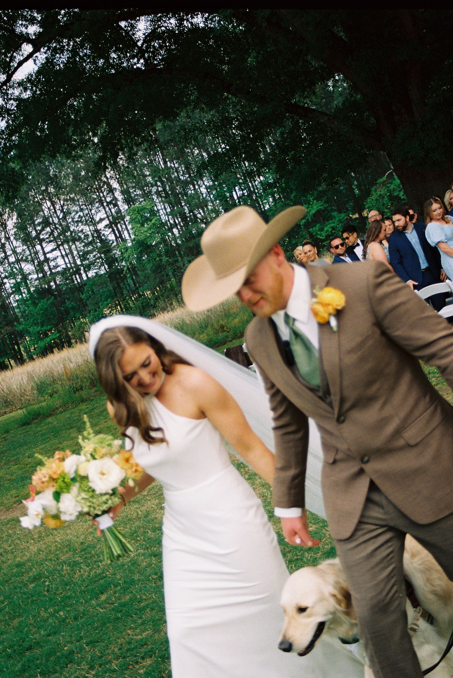 Bride and groom walking down outdoor aisle with dog, groom in tan cowboy hat and brown suit, bride holding colorful bouquet.
