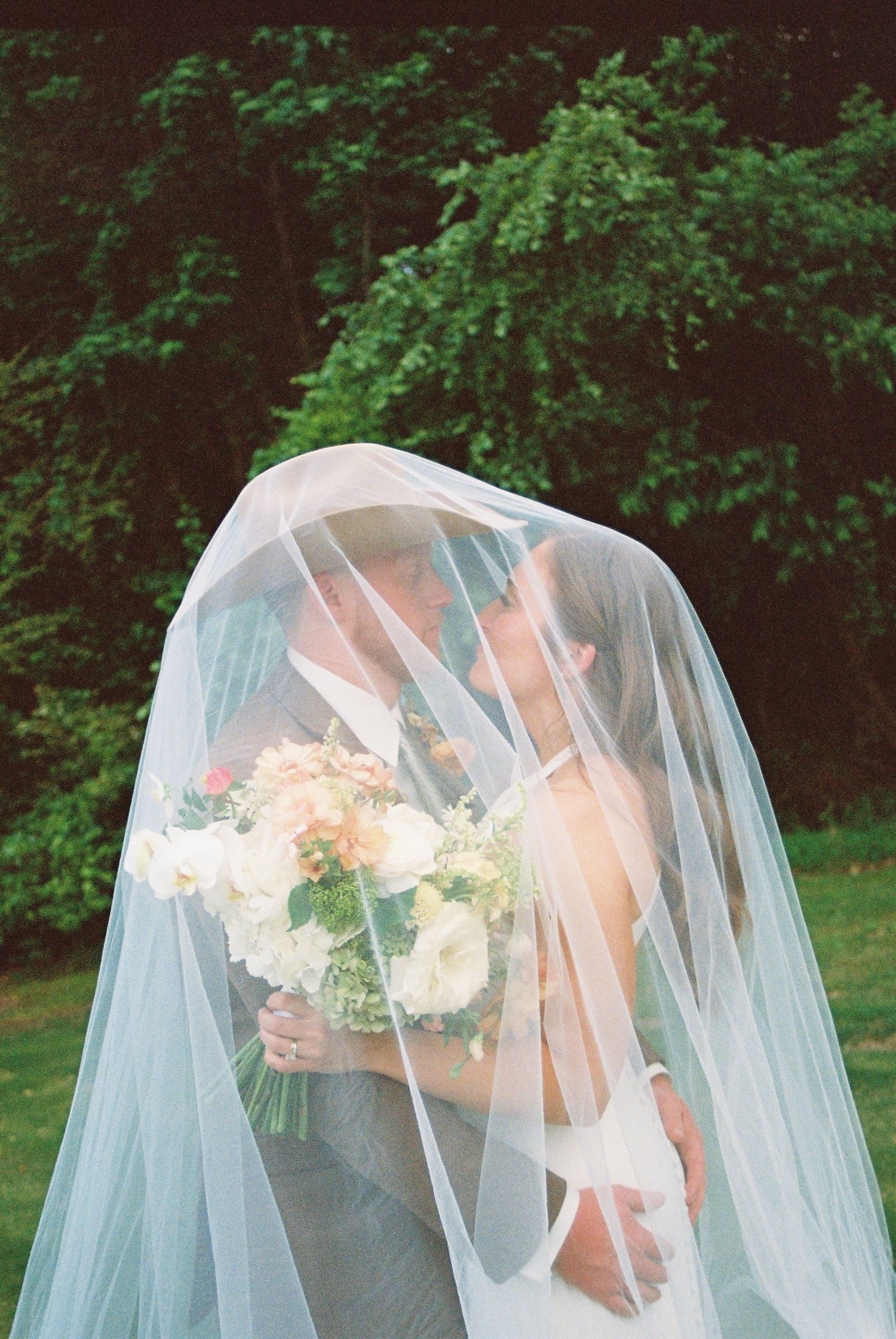 Bride and groom kissing under a flowing white veil, bride holding a pastel bouquet, with lush green trees in the background.