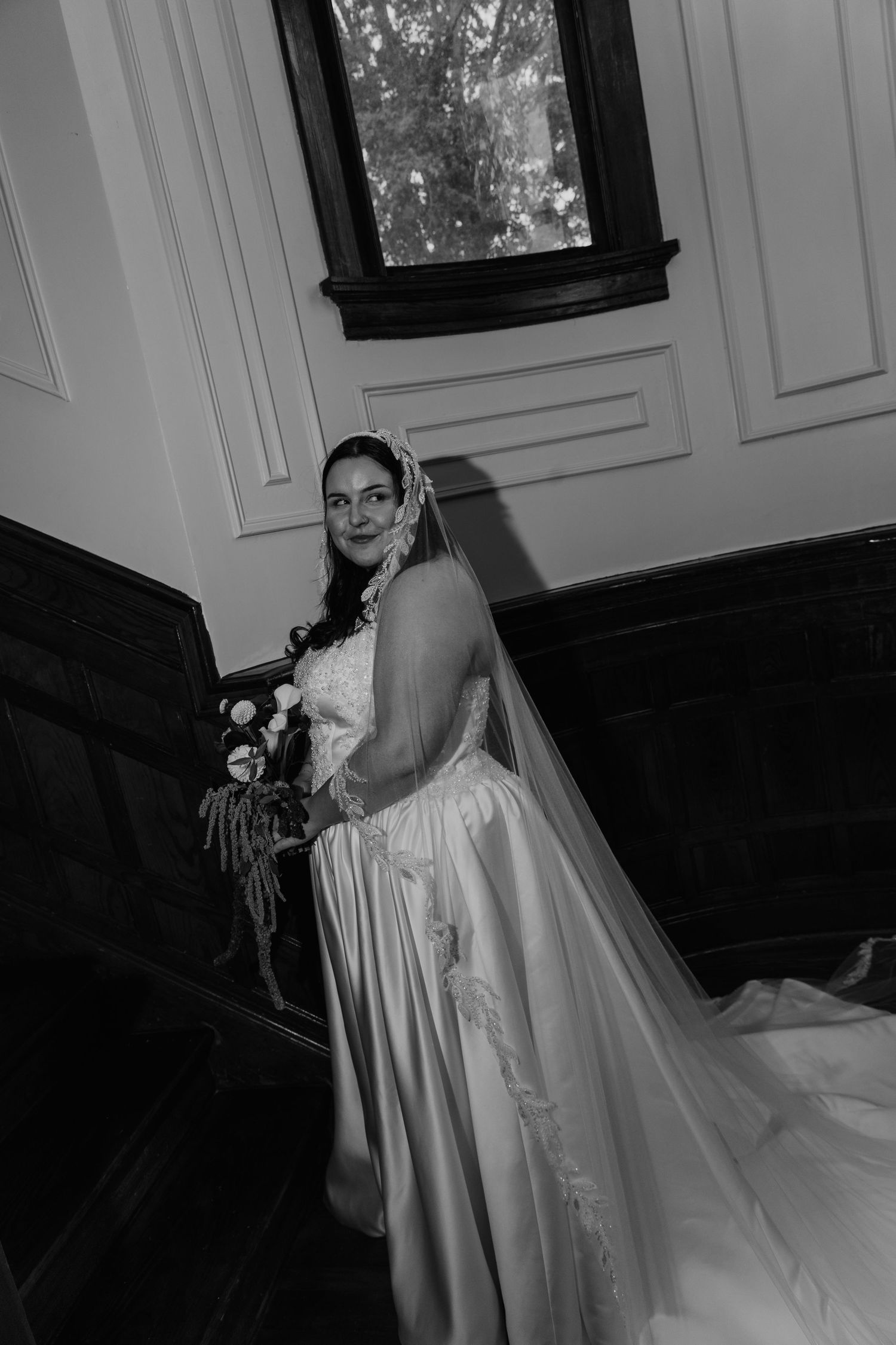 Black and white photo of a bride in a satin wedding gown and lace-trimmed veil holding a bouquet on a dark wooden staircase.