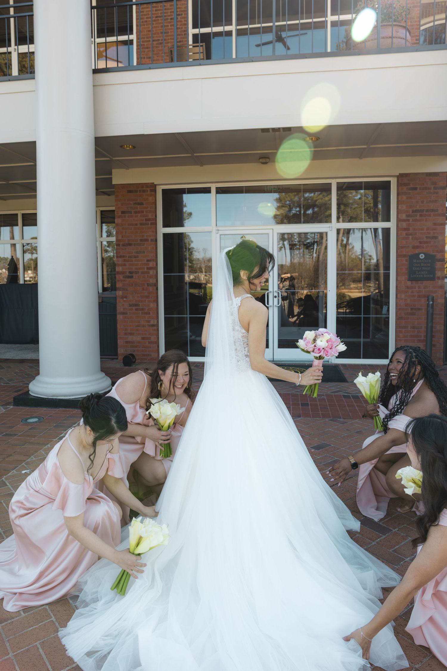 Bride in white gown with bridesmaids in blush pink dresses arranging her train outside a brick venue.