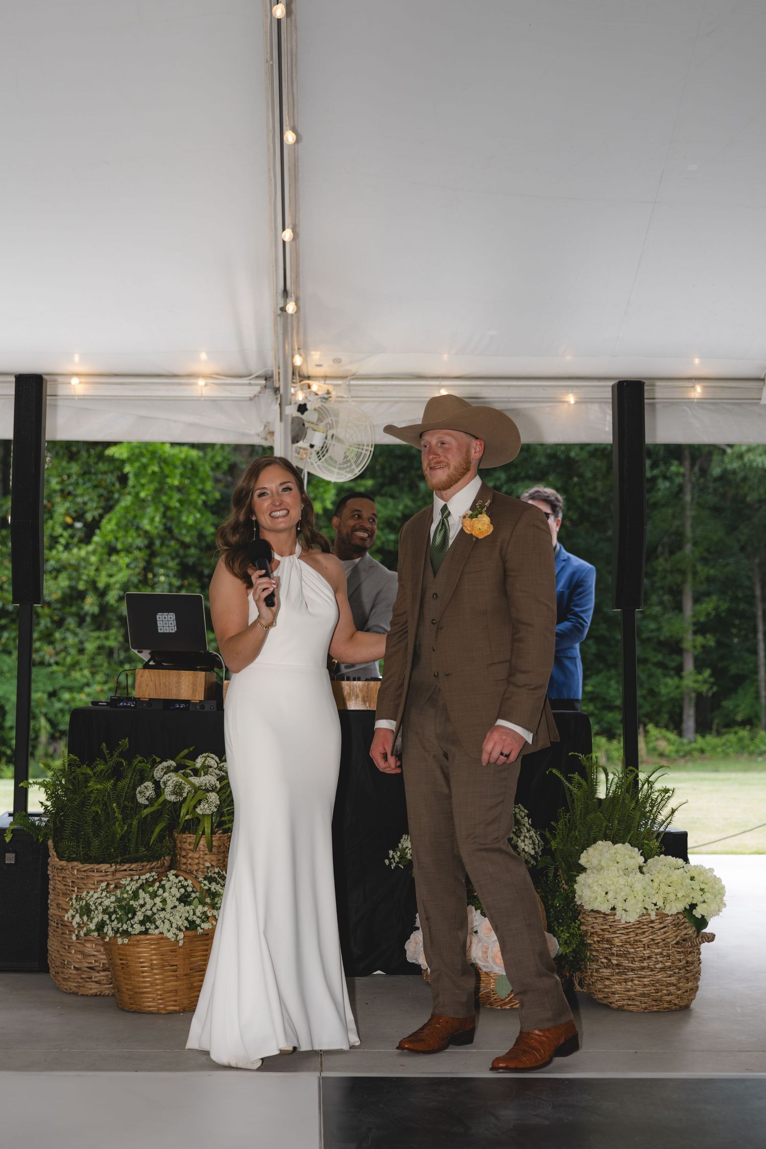 Smiling bride and groom in cowboy hat make announcements with microphone at their wedding reception.