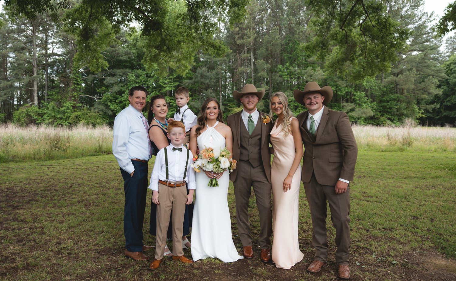 Wedding party family portrait with bride in white gown and groom in brown suit and cowboy hat outdoors.