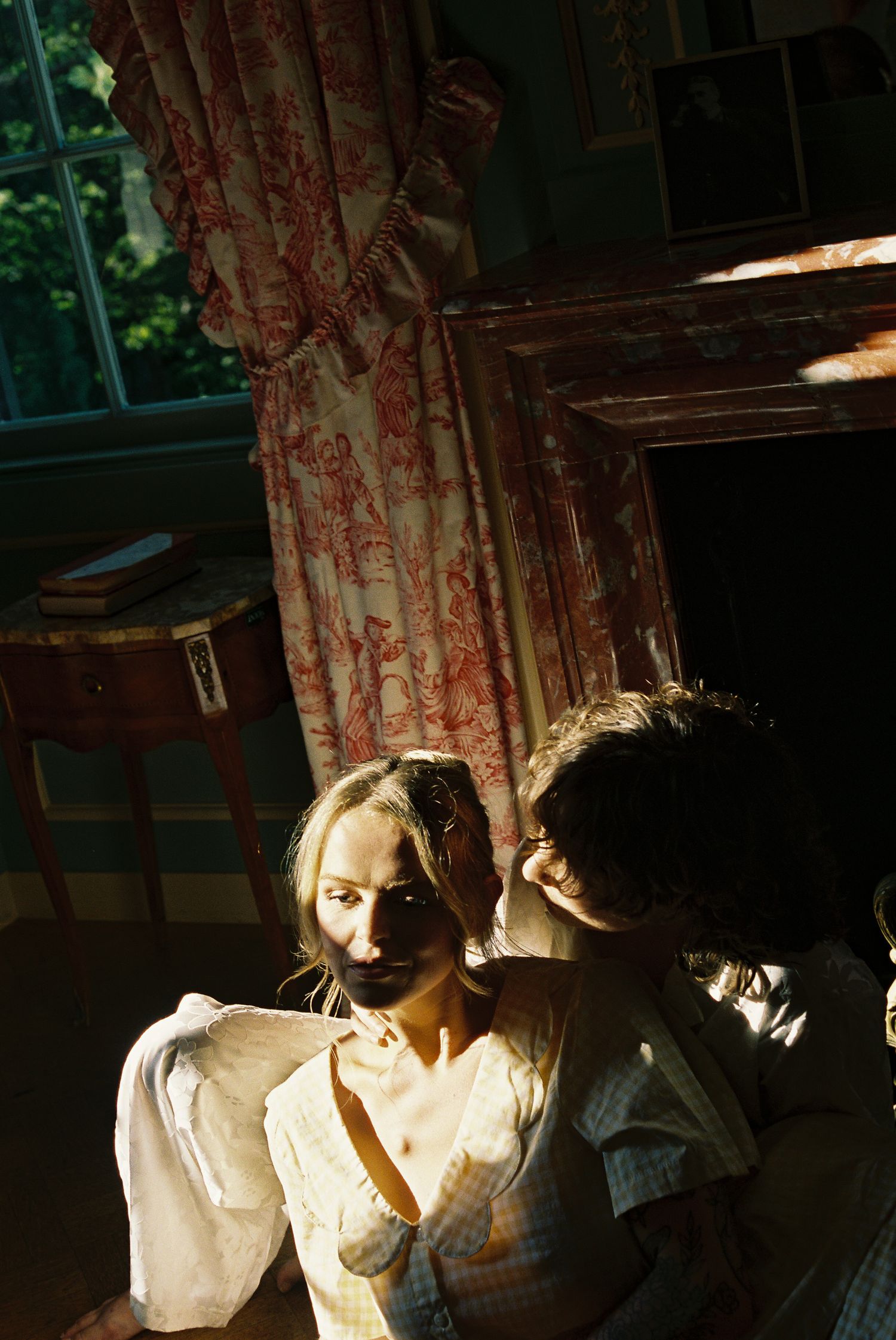Two women in vintage white dresses stand in a sunlit room with red floral curtains and antique furniture.