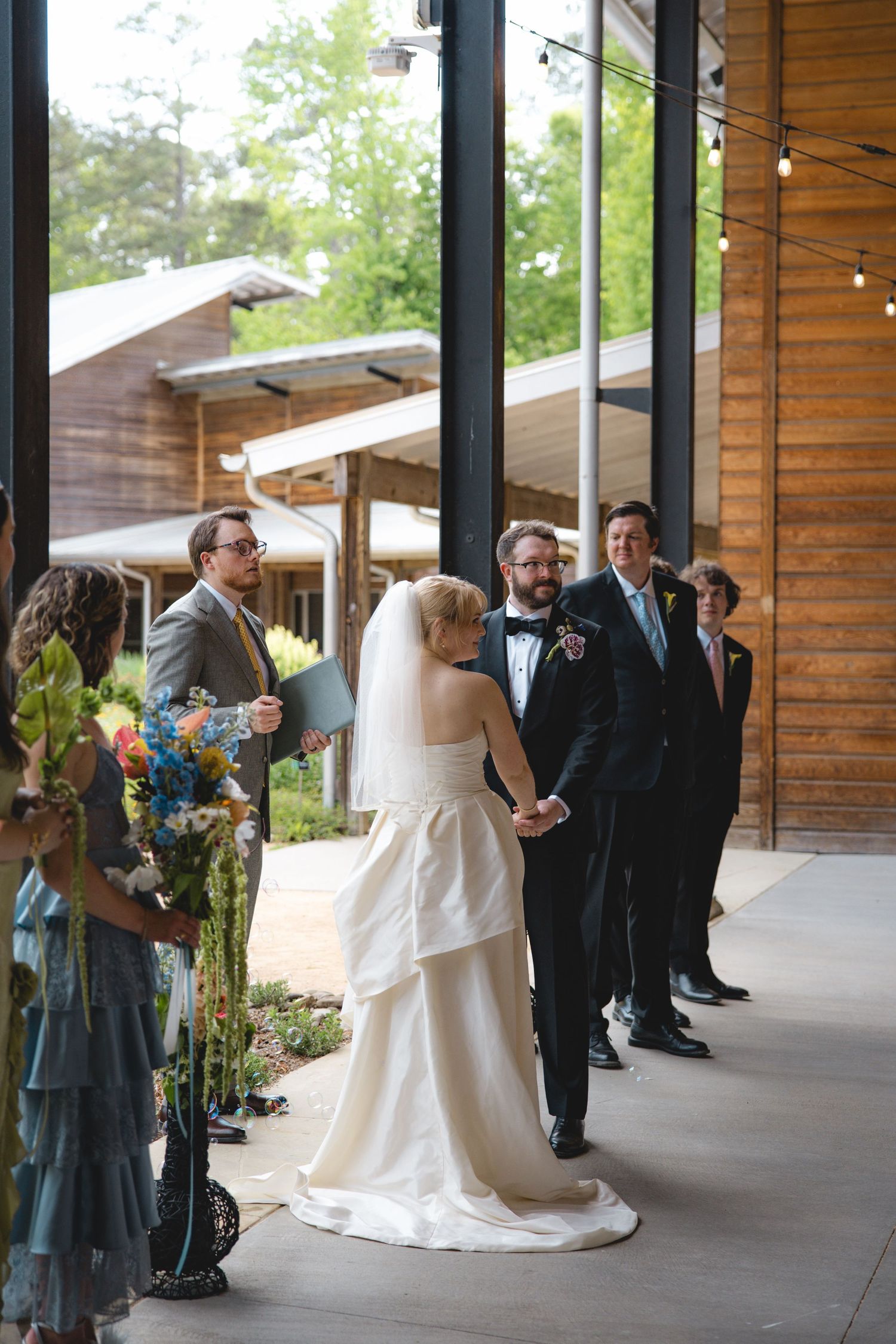 Bride and groom hold hands at outdoor wedding ceremony in rustic wood venue with groomsmen and officiant.