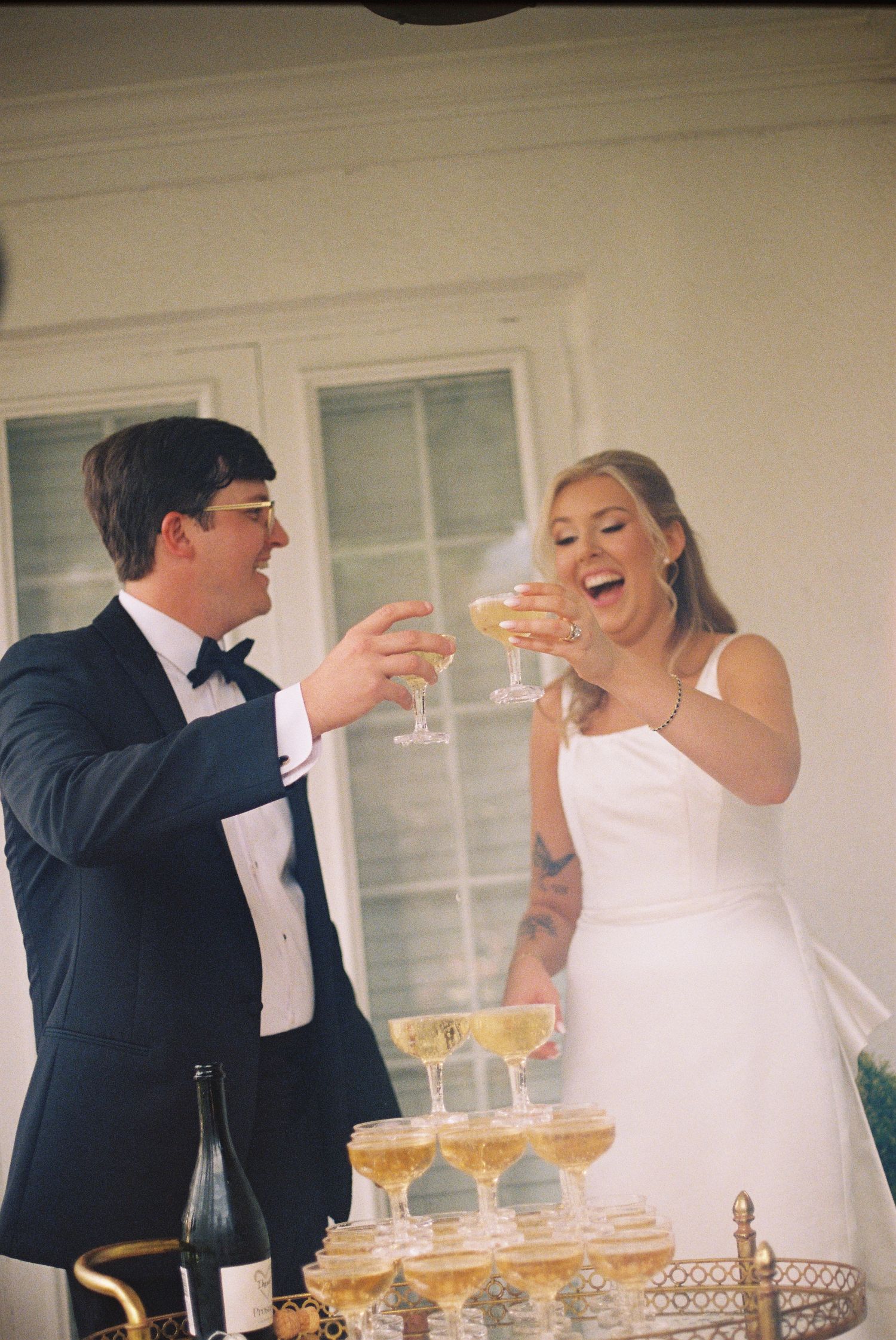 Bride and groom laughing while toasting champagne glasses at a wedding reception with a champagne tower display.