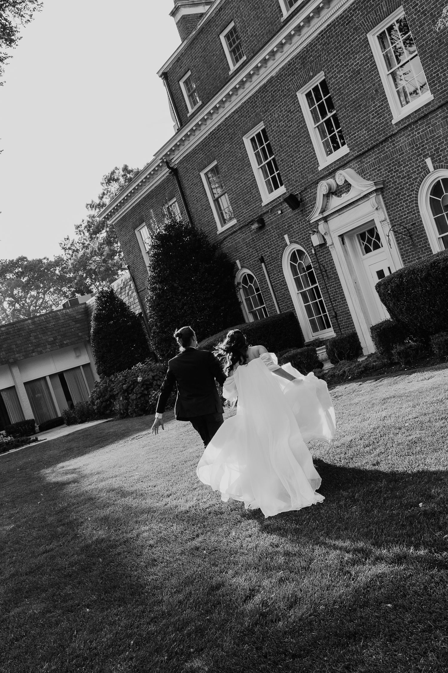Bride and groom running hand in hand toward a grand brick manor in a romantic black and white wedding photo.