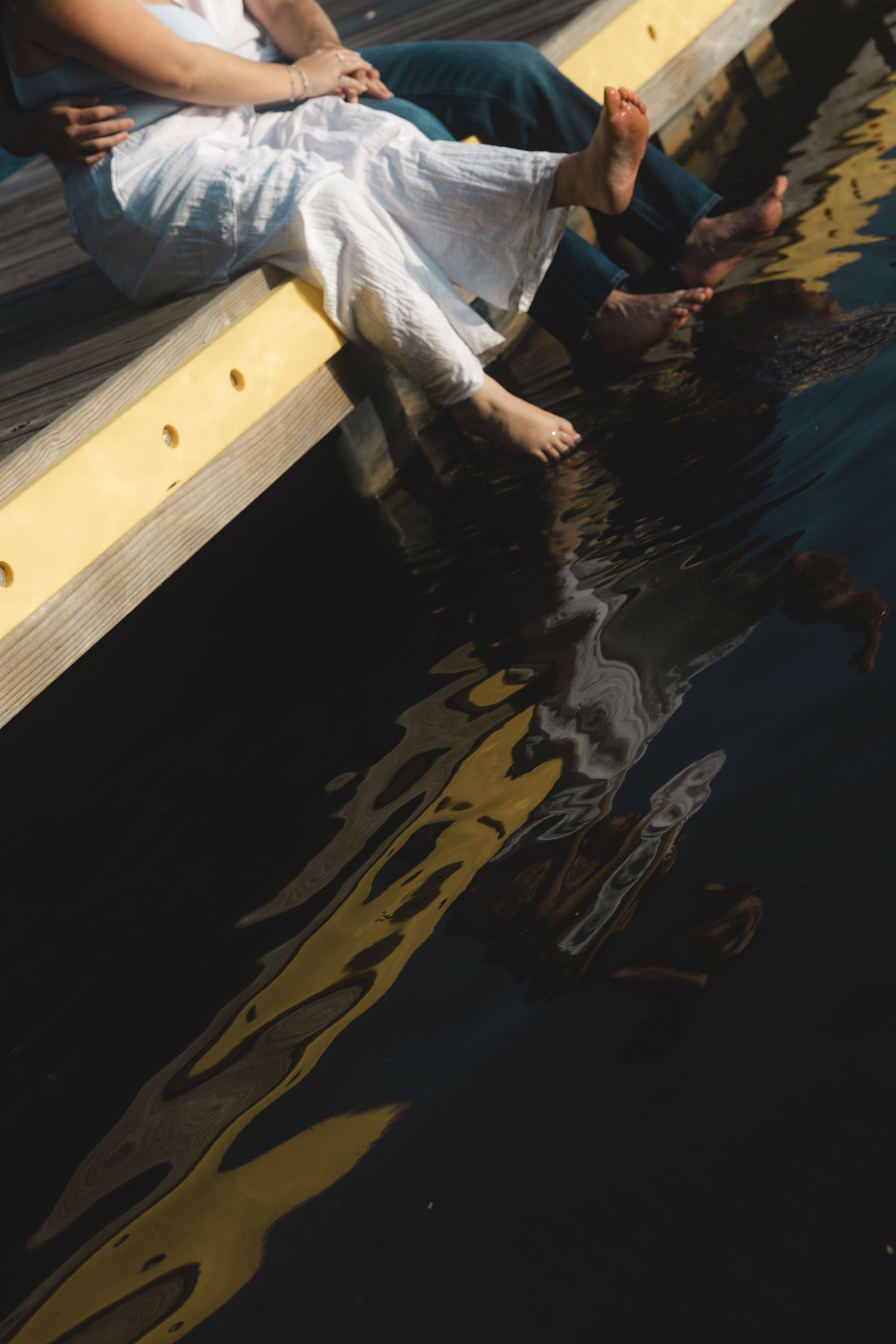 Couple sitting on wooden dock with bare feet dangling over water, reflections visible below.