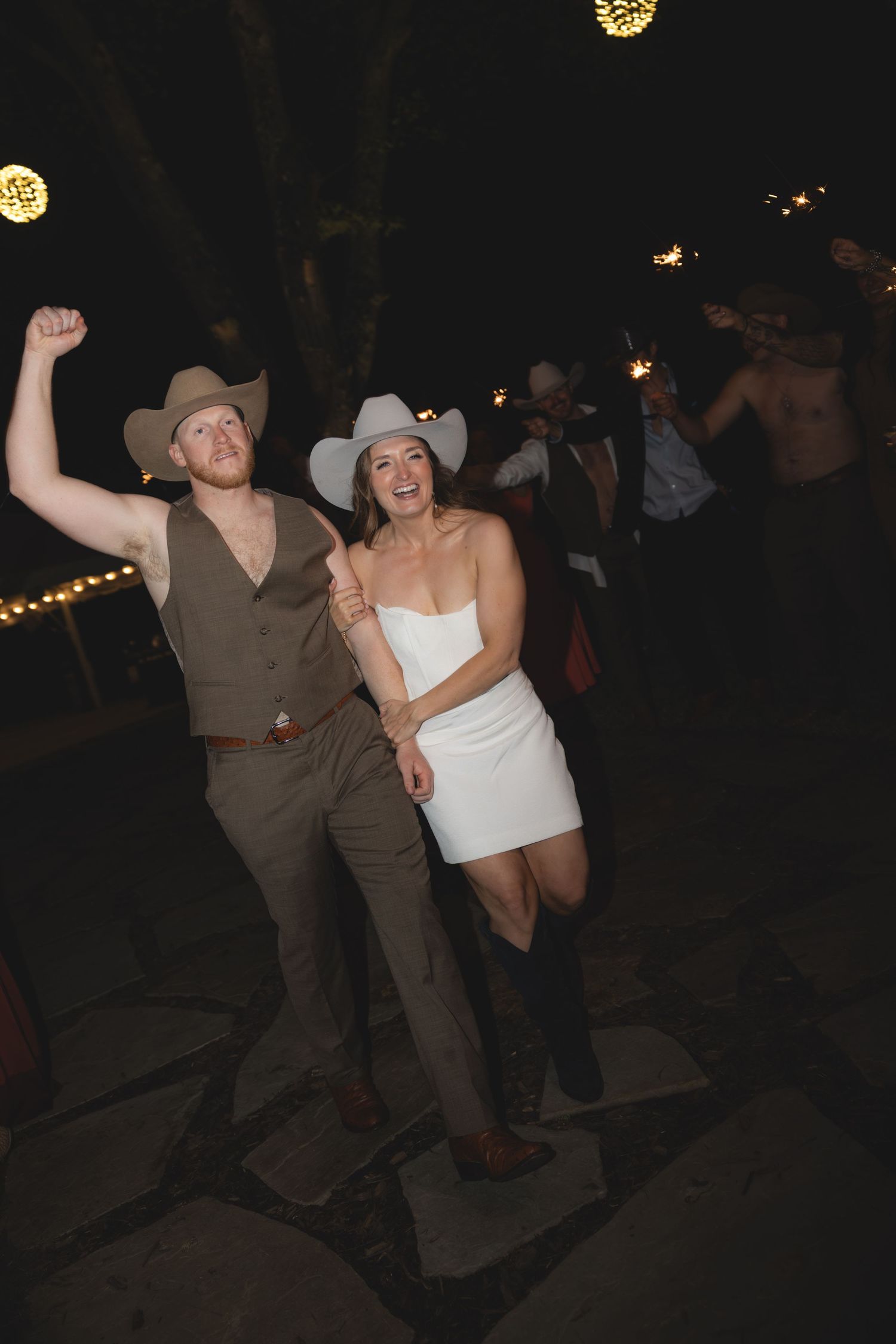 Smiling bride and groom in cowboy hats celebrating and dancing together outdoors at their wedding reception at night.