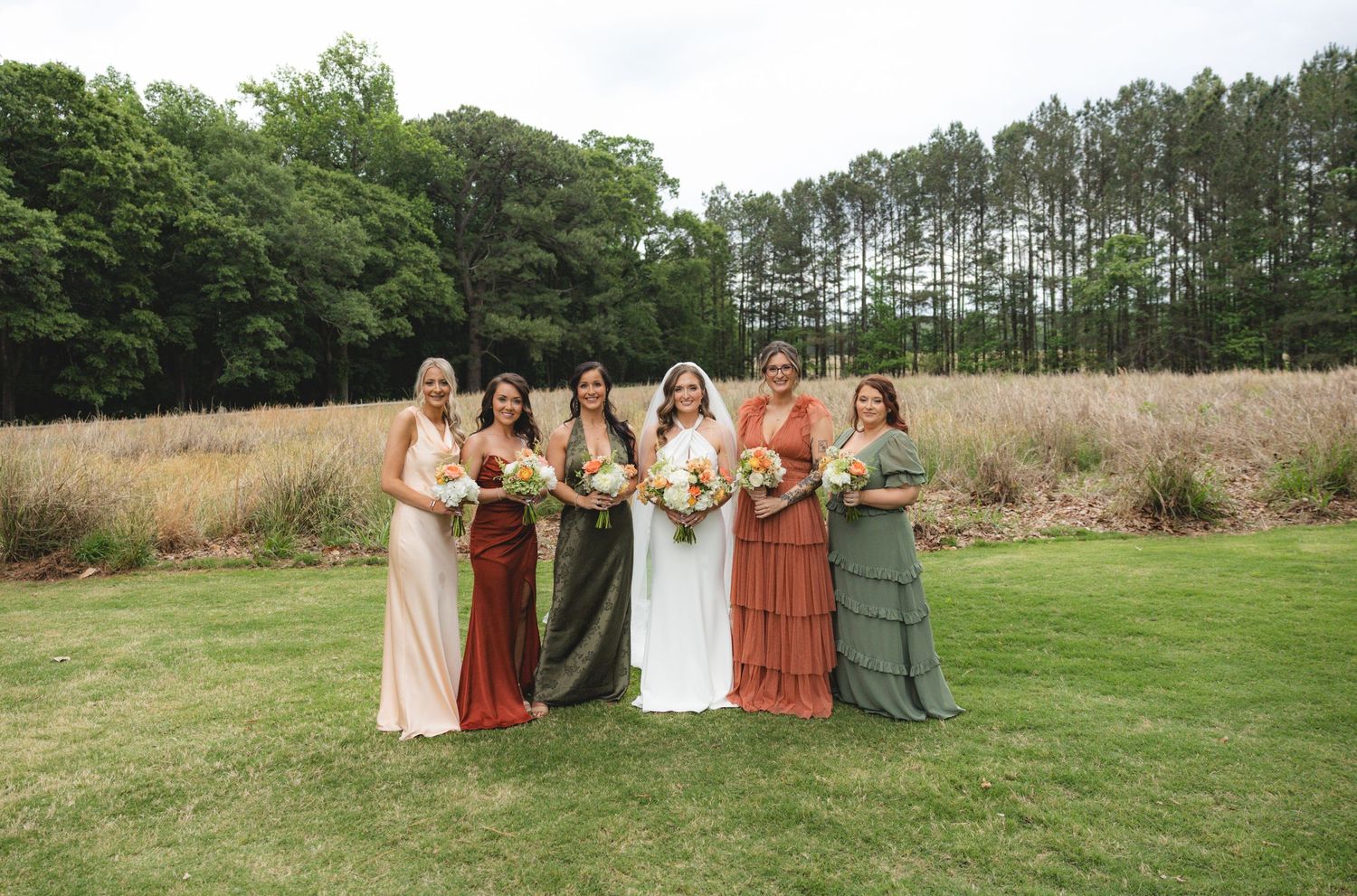 Bride in white gown poses with five bridesmaids in mismatched earth-tone dresses holding bouquets outdoors.