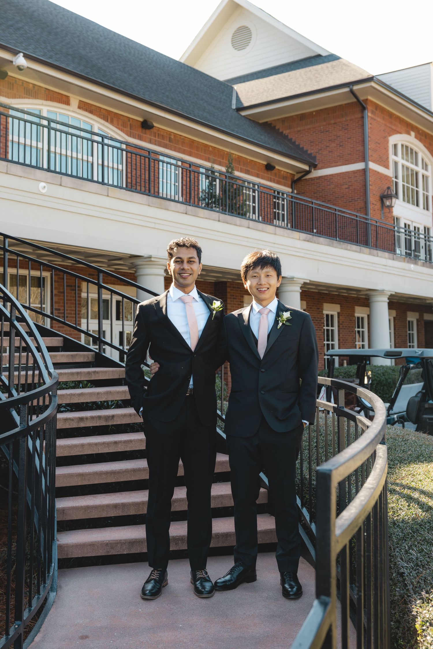 Two groomsmen in black suits with white ties posing on staircase outside brick wedding venue.