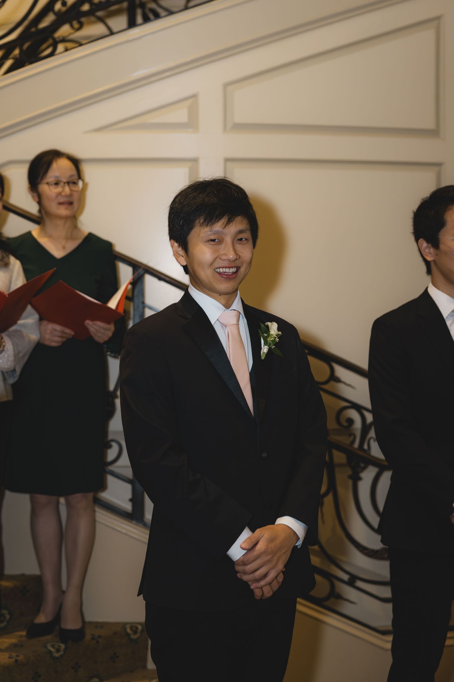 Smiling groom in black suit with pink tie awaits bride at wedding ceremony altar.