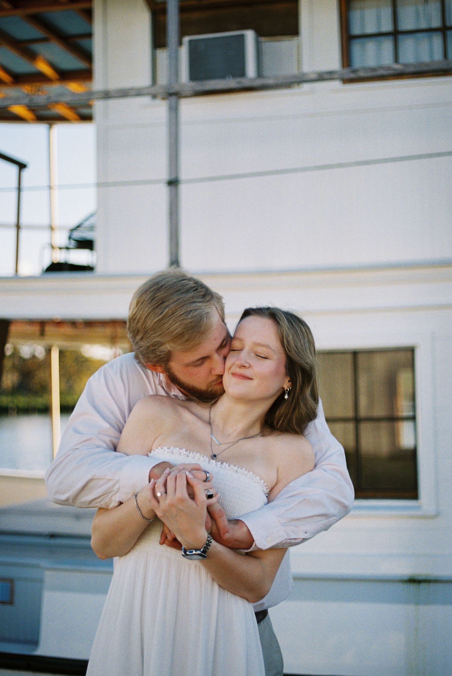 Romantic couple embracing outdoors, man kissing woman's cheek while she smiles, wearing white off-shoulder dress near white building.
