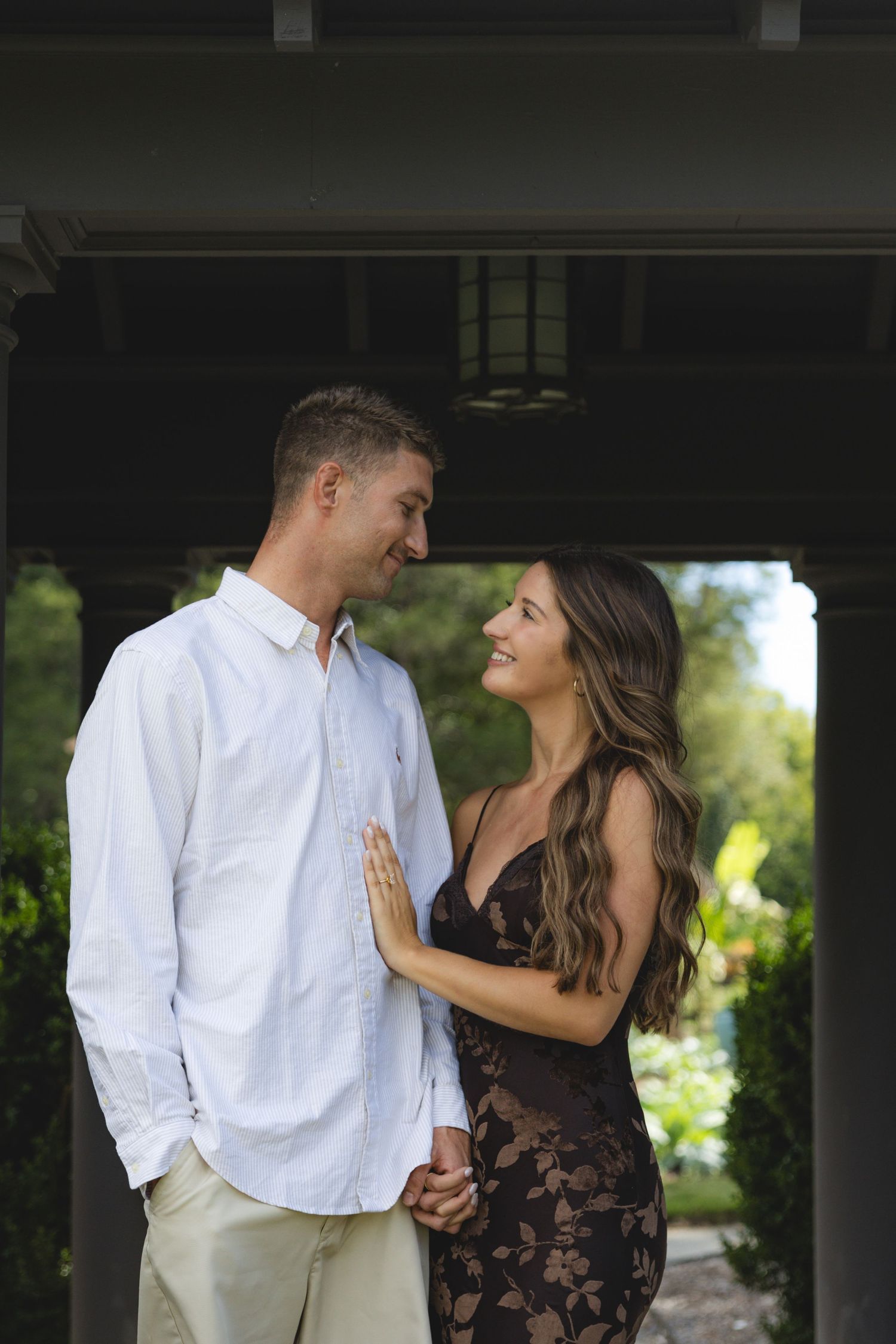 A smiling couple holds hands and gazes at each other outdoors, man in white shirt, woman in floral dress.