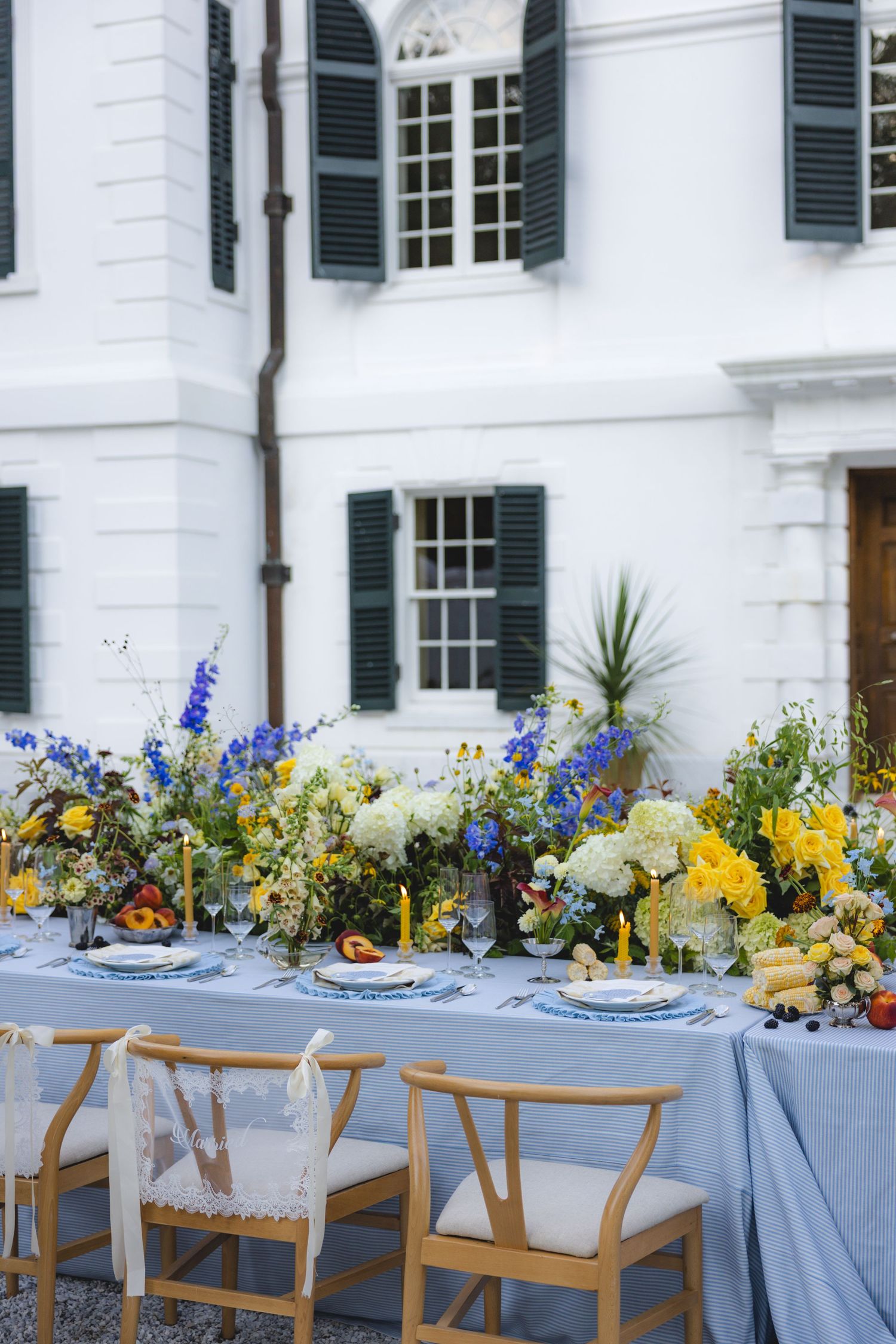Elegant outdoor wedding reception table with blue linen, yellow candles, and vibrant floral arrangements before a white manor.