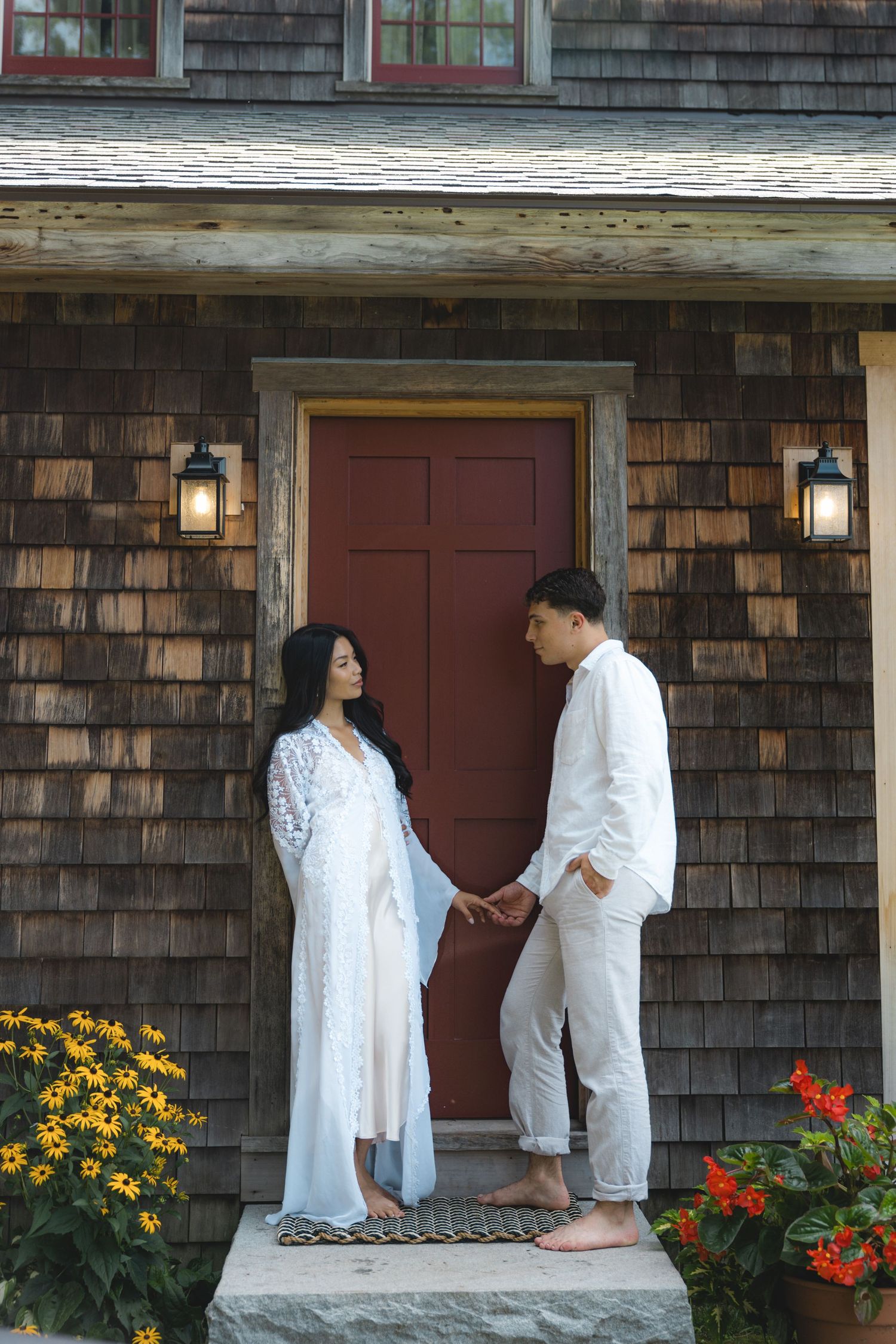 Couple in white outfits holding hands on porch of rustic cedar shingle home with red door and flowers.