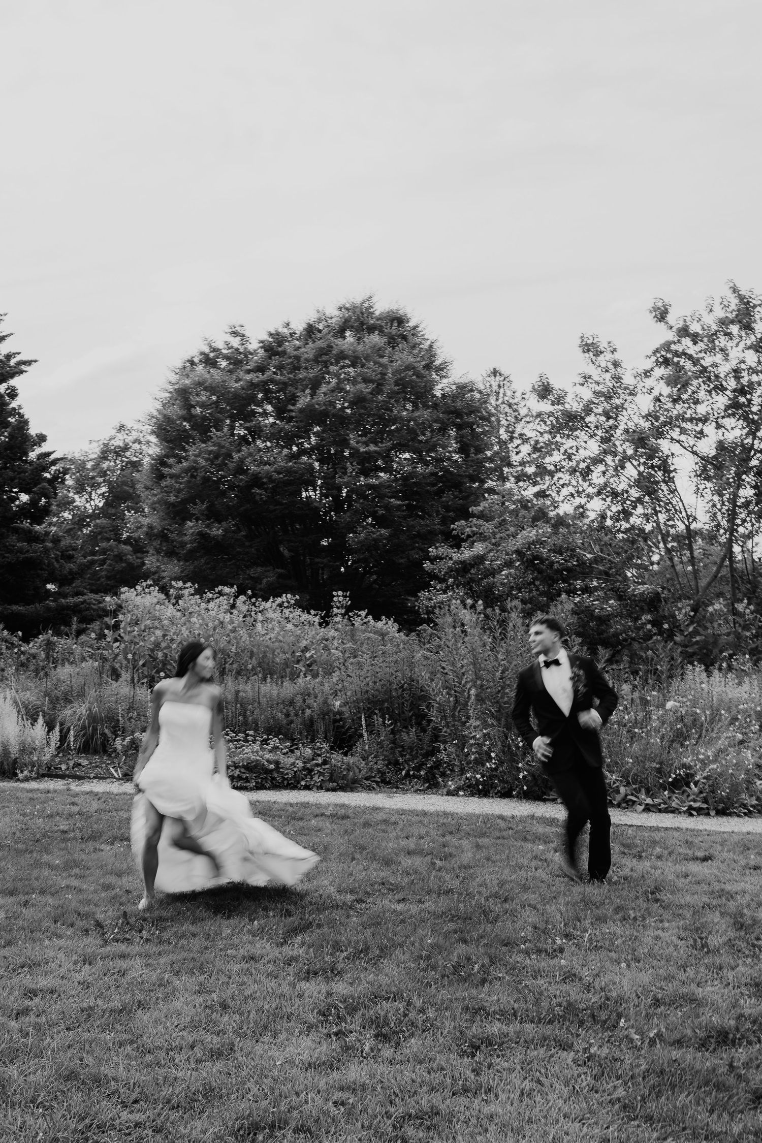 Black and white photo of a bride and groom joyfully running across a garden lawn on their wedding day.