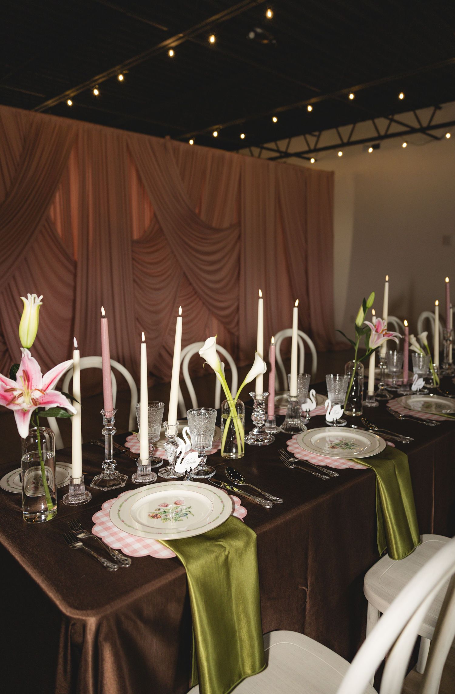 Elegant wedding reception table with green velvet runner, pink lilies, white taper candles, and draped brown backdrop.