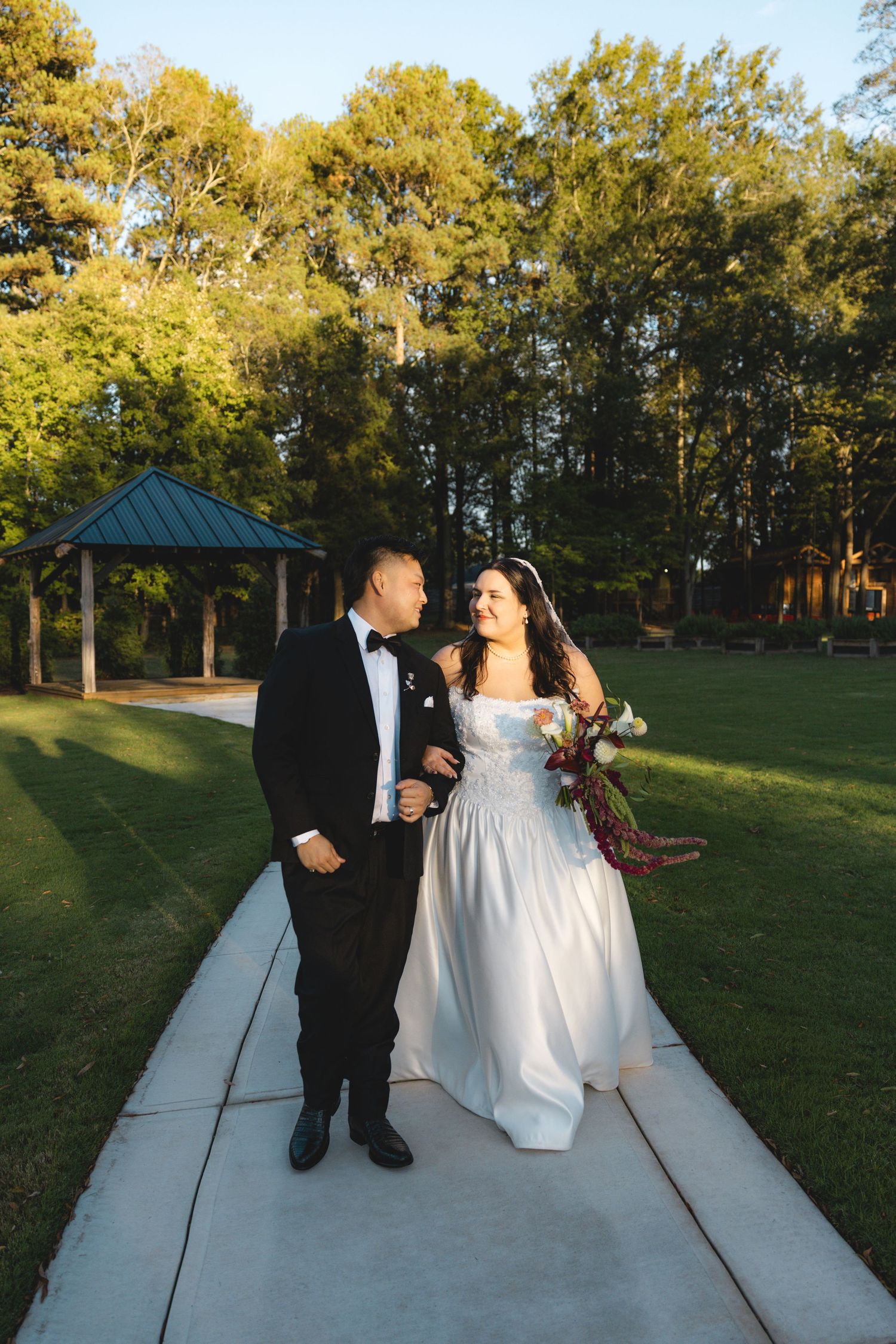Smiling bride in a white gown holding a bouquet walks arm in arm with a groom in a black tuxedo on a garden path near a gazebo.