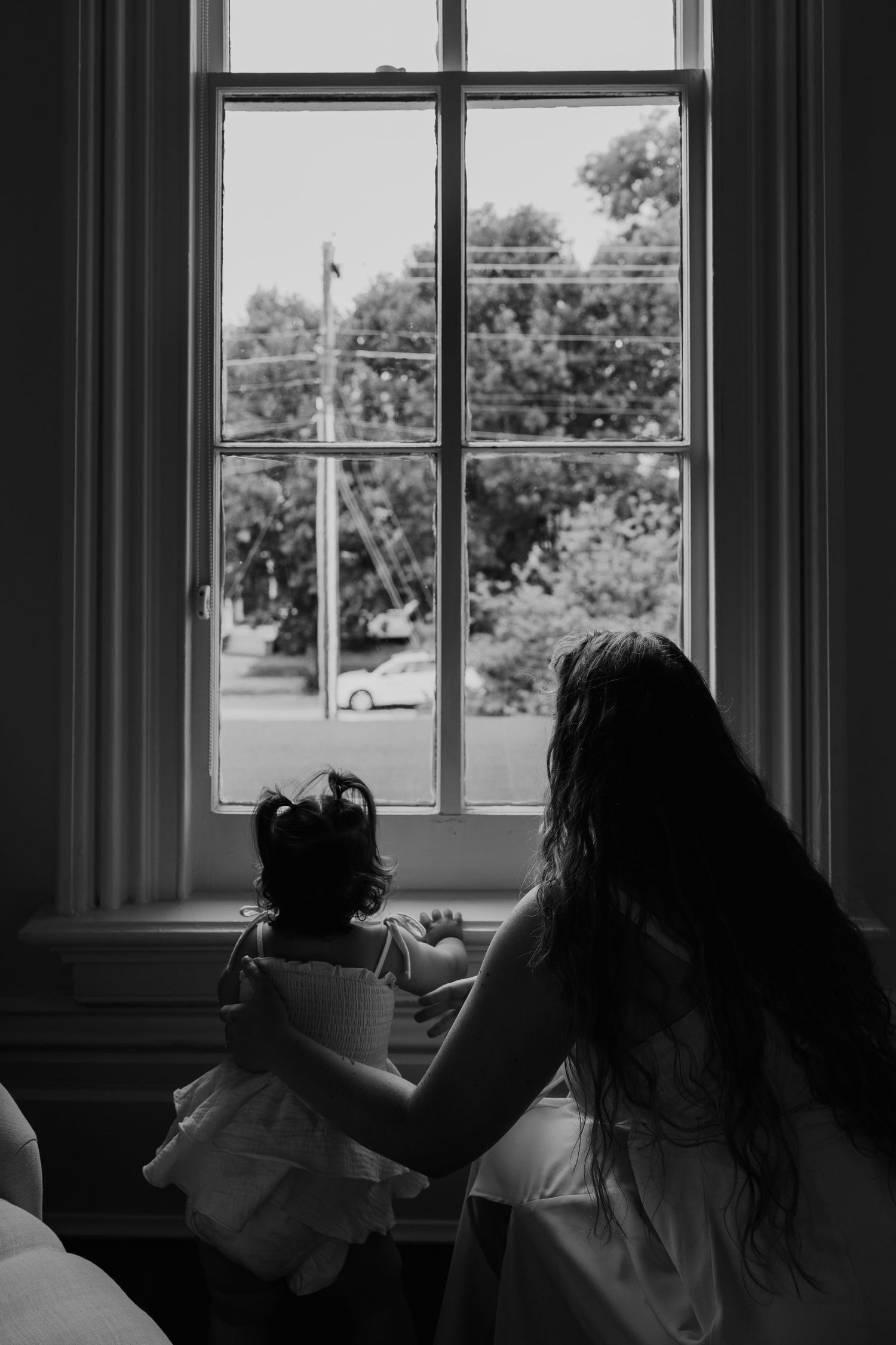 Black and white photo of a mother and young daughter looking out a large window together.