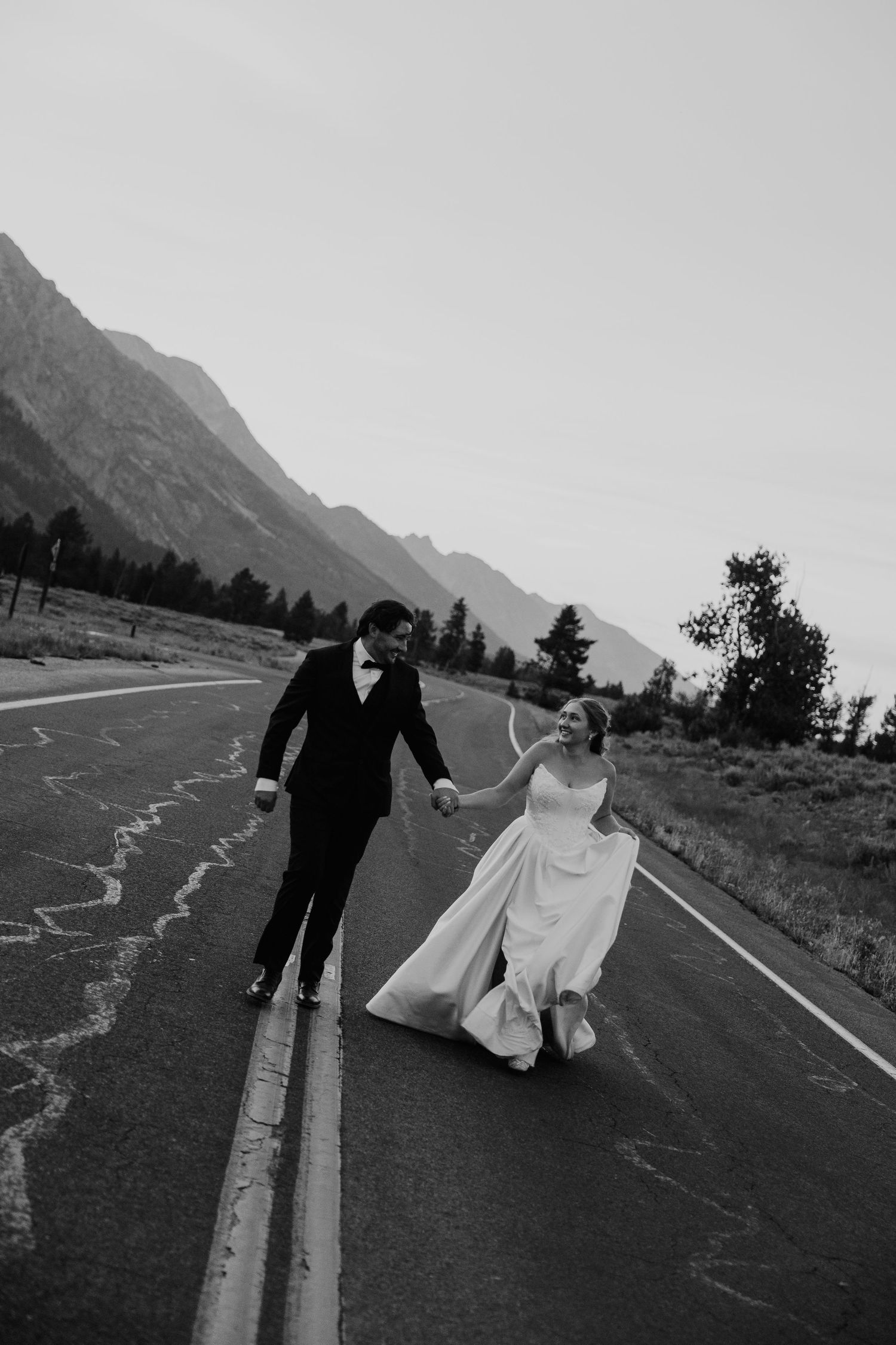 Black and white photo of a bride and groom holding hands walking on a mountain road in wedding attire.