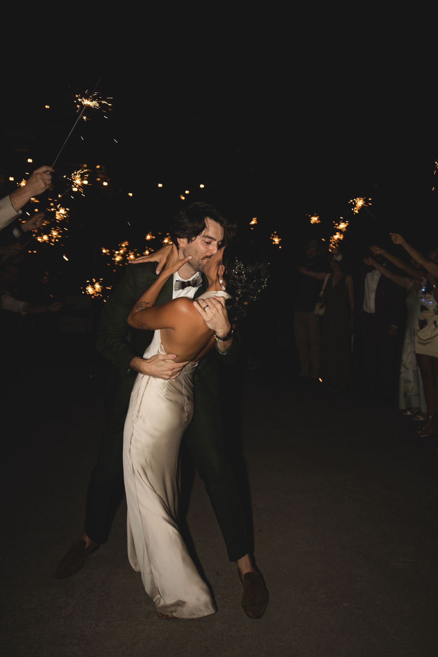 A newlywed couple shares a romantic kiss surrounded by guests holding sparklers at their night wedding reception.