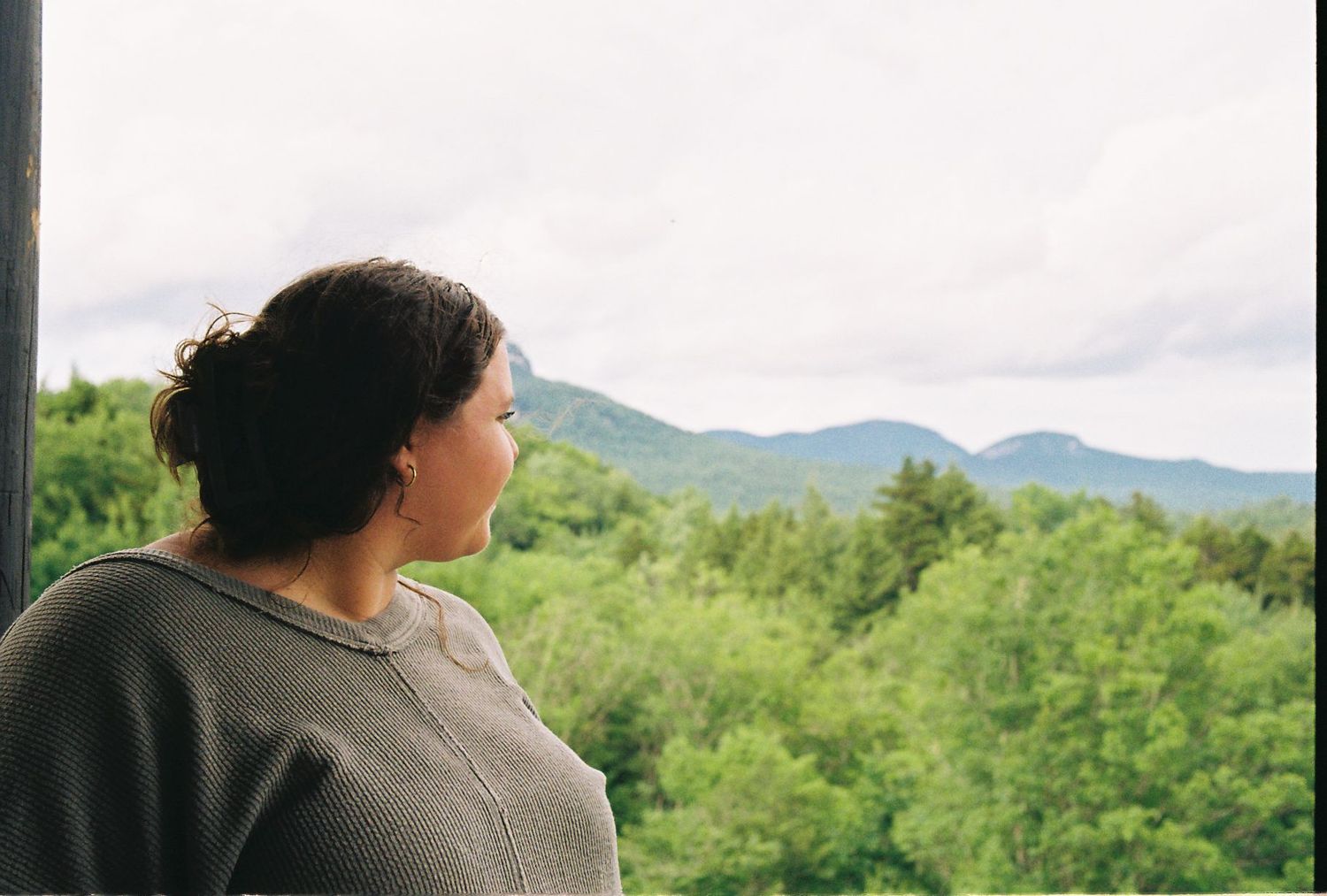 Woman in gray sweater gazes at lush green mountain landscape from elevated viewpoint on cloudy day.