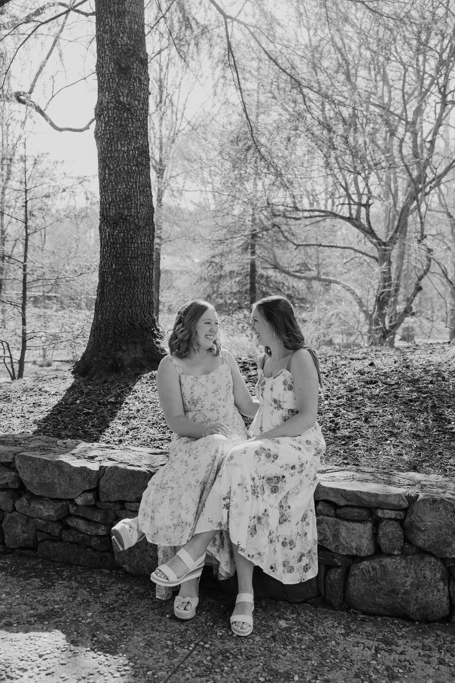 Two young women in floral dresses sit on a stone wall laughing together in a black and white outdoor photo.