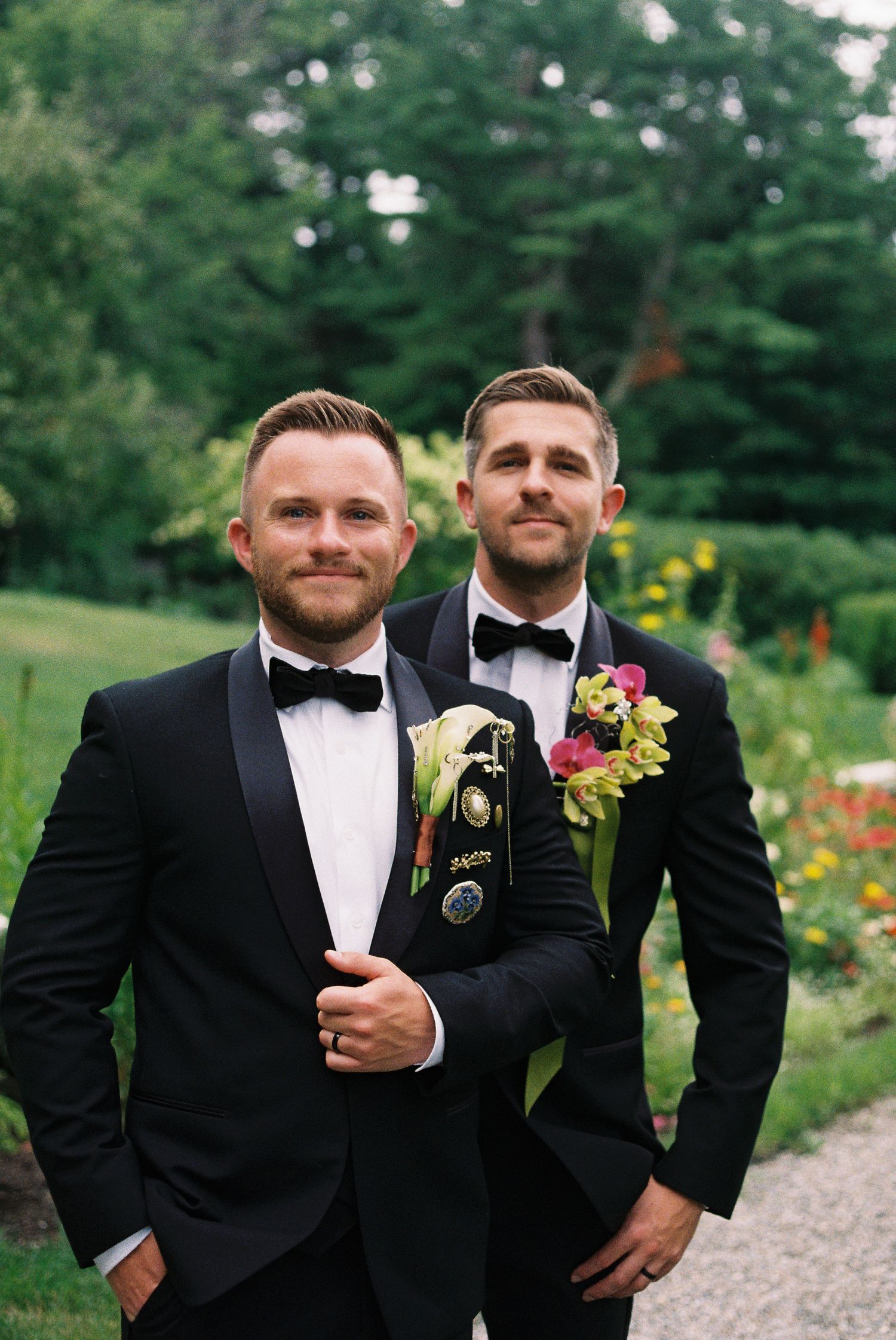 Two grooms in black tuxedos with bow ties and floral boutonnieres pose outdoors in a garden setting.