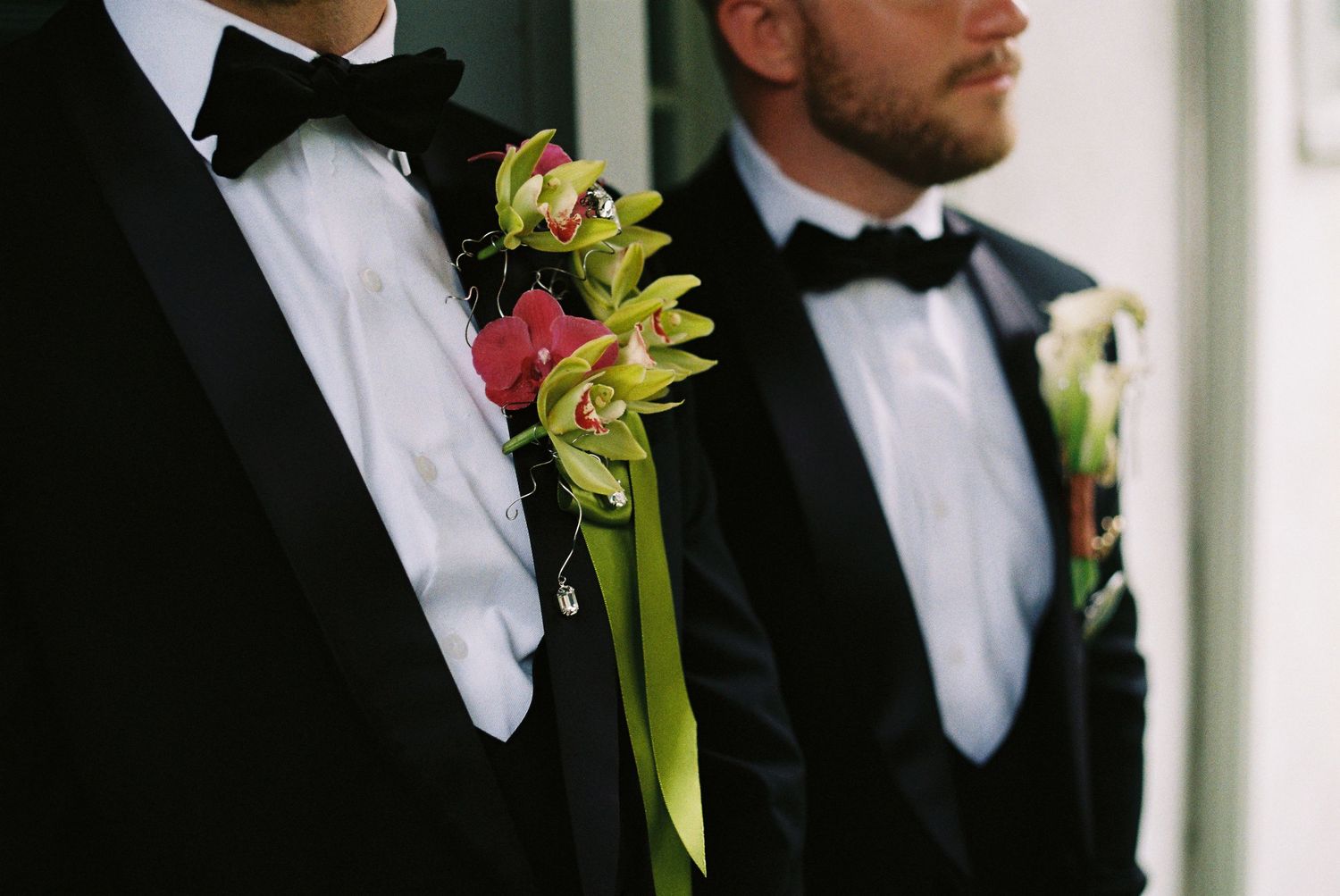 Two grooms in black tuxedos with bow ties wearing floral boutonnieres with green orchids at a wedding.