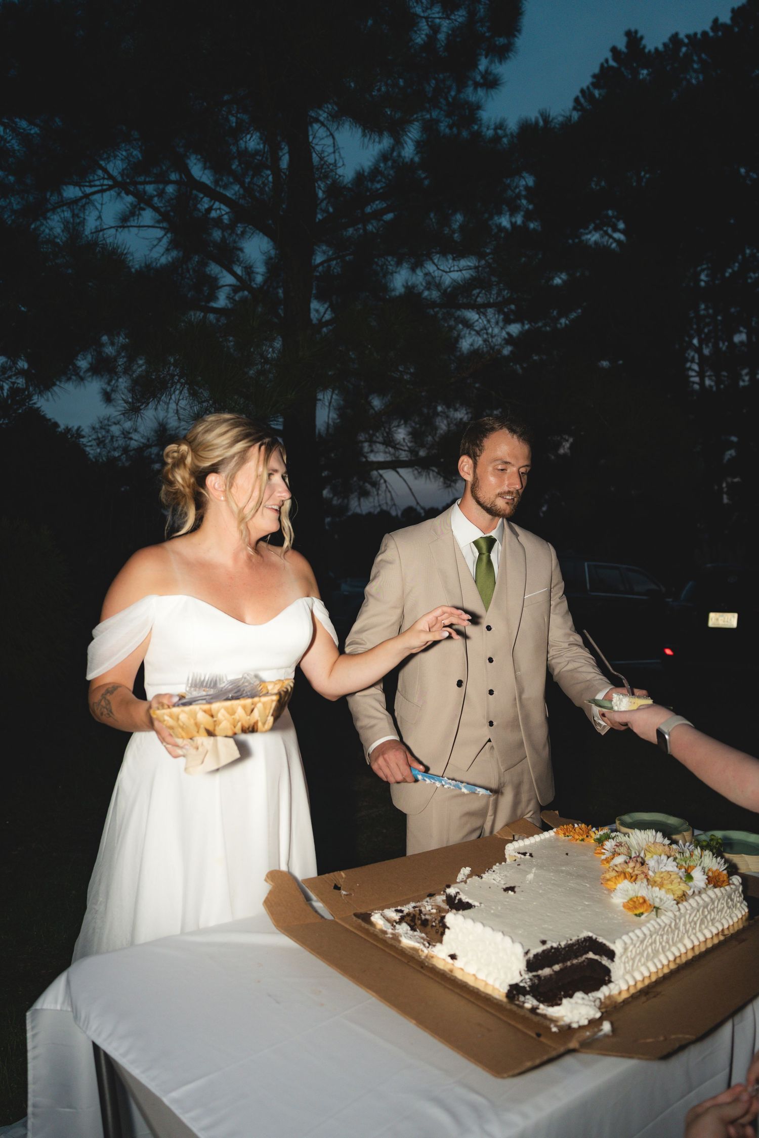 Bride and groom cutting their wedding cake outdoors at night, bride laughing while holding a plate of food.