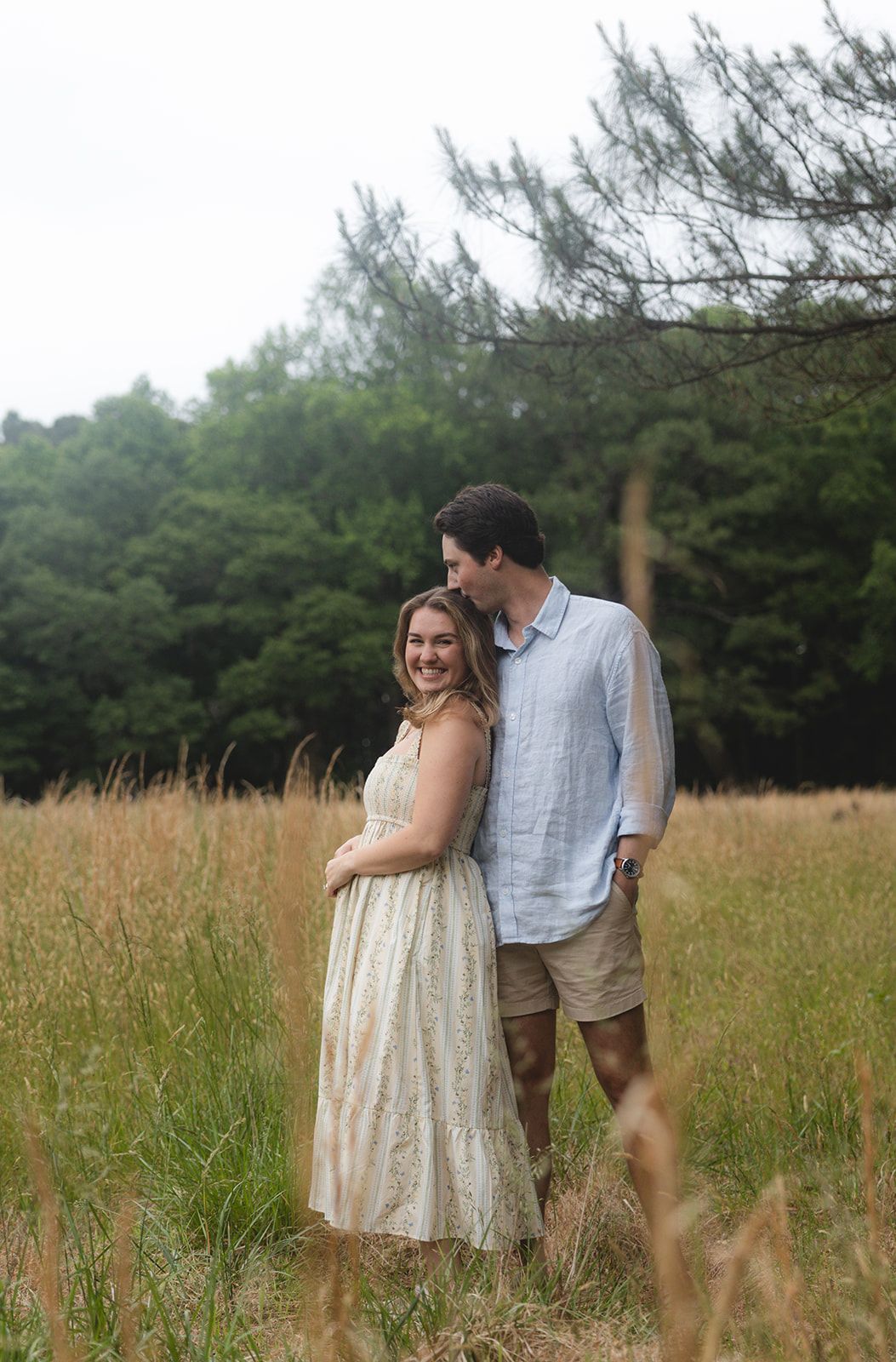 A smiling couple stands embracing in a golden meadow with lush green trees in the background.
