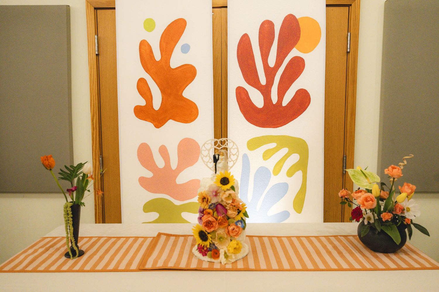 Colorful Matisse-inspired art panels backdrop a floral cake on striped tablecloth with vibrant flower arrangements.