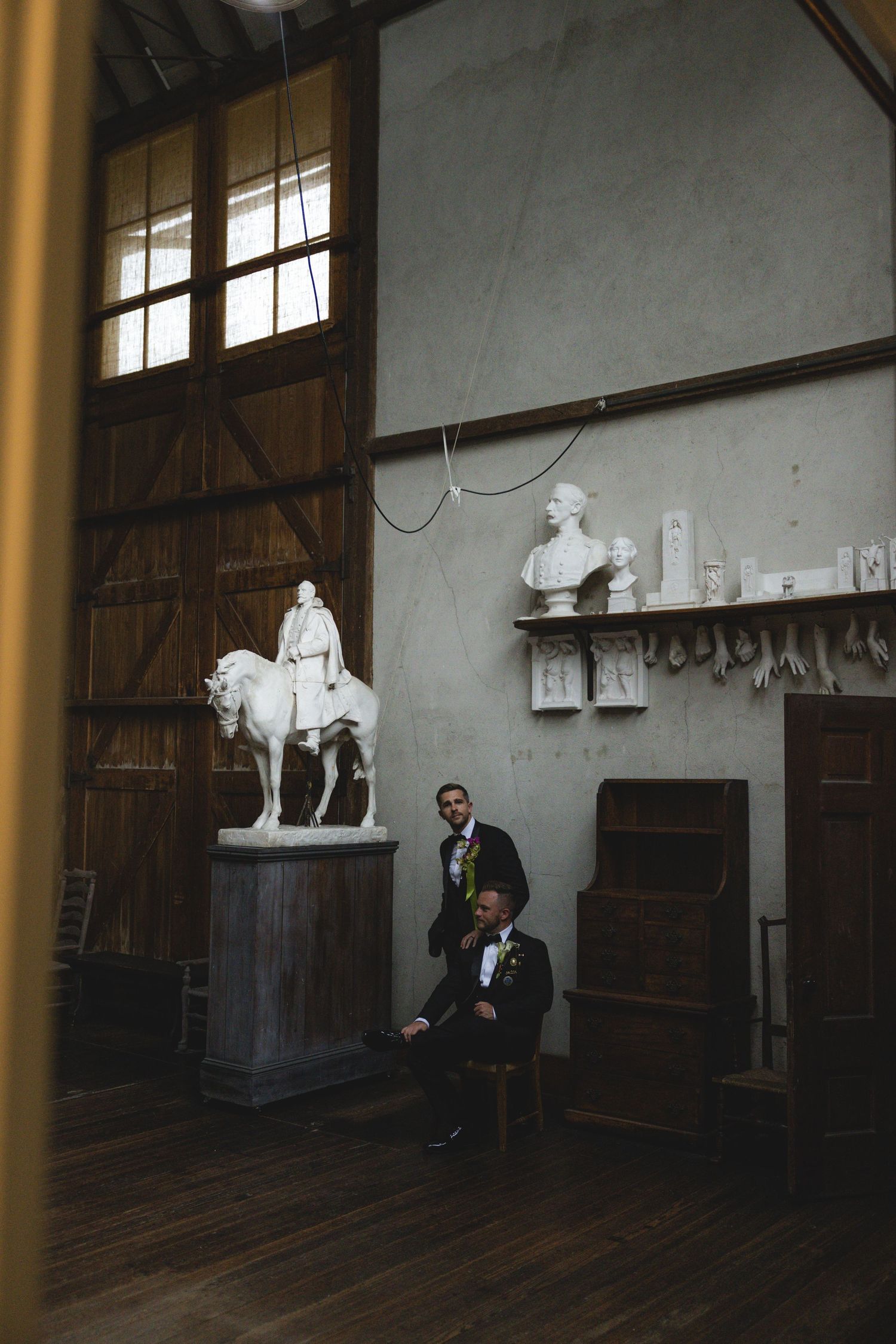 Two grooms in tuxedos pose in a grand hall with white marble sculptures and high wooden ceilings.