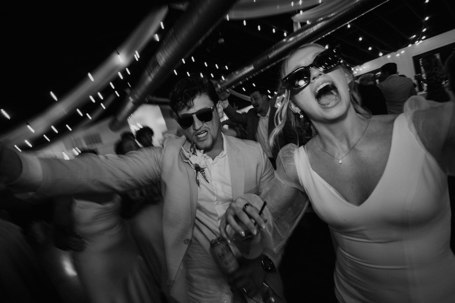 Black and white photo of joyful couple dancing at wedding reception wearing sunglasses under string lights.