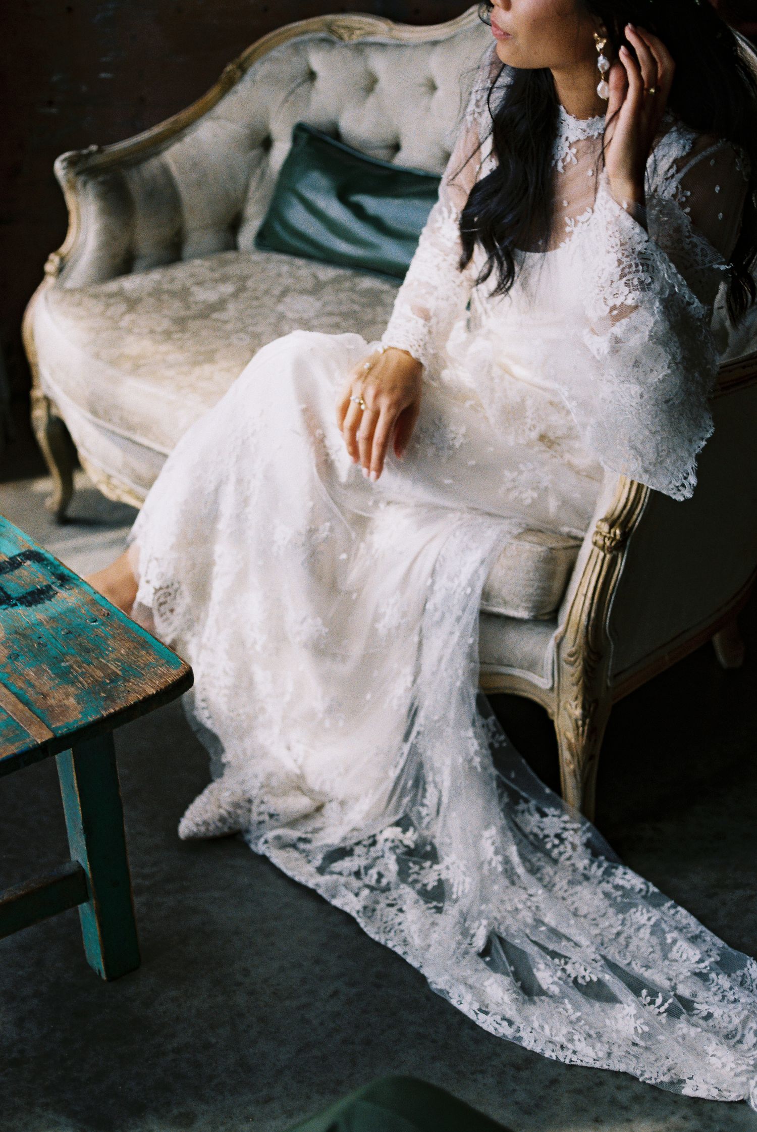 Bride in long-sleeve lace wedding gown sitting elegantly on vintage tufted sofa with teal accent pillow.