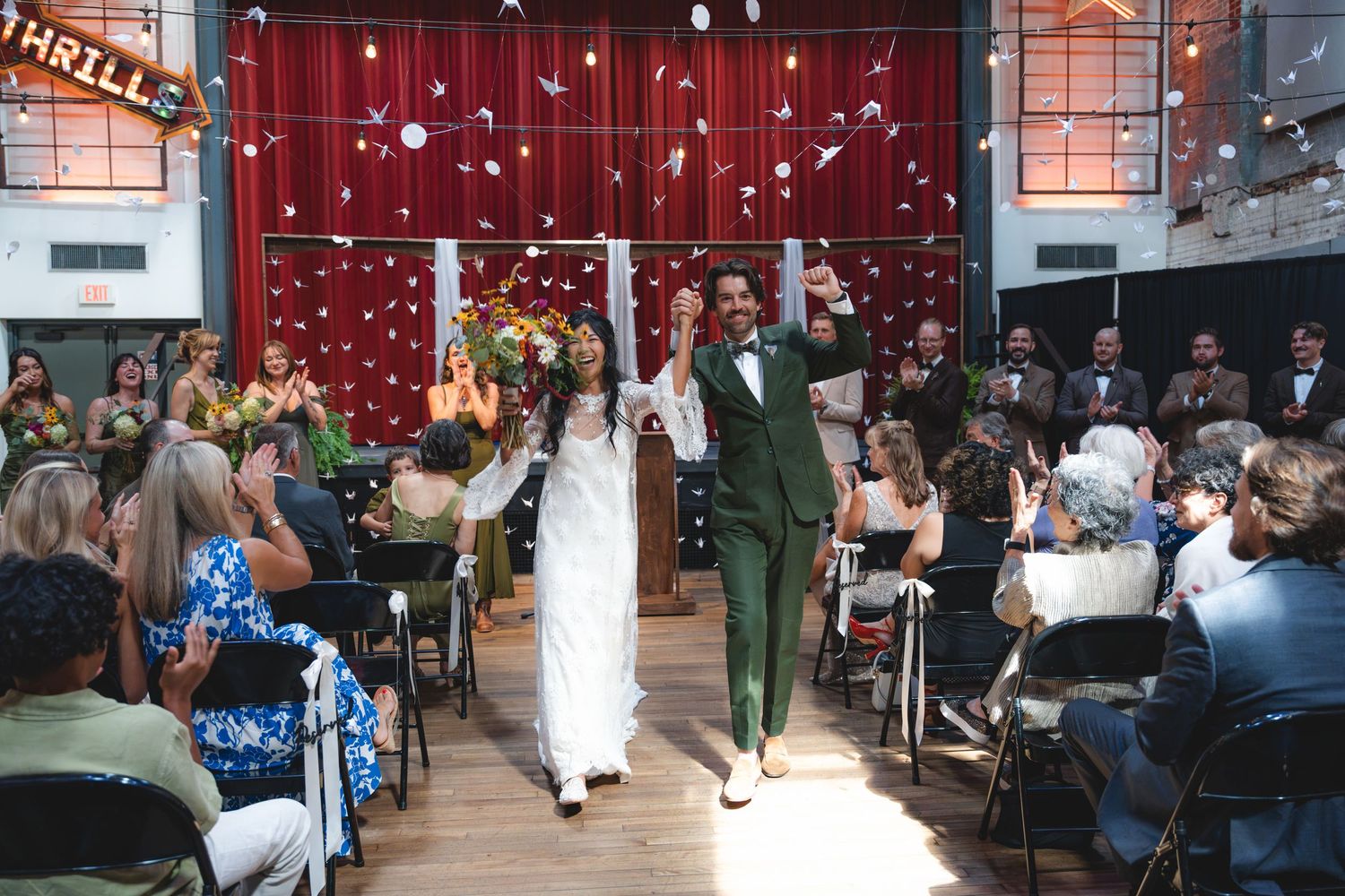 Newlyweds walk down the aisle amid confetti at a festive indoor wedding ceremony with guests applauding.