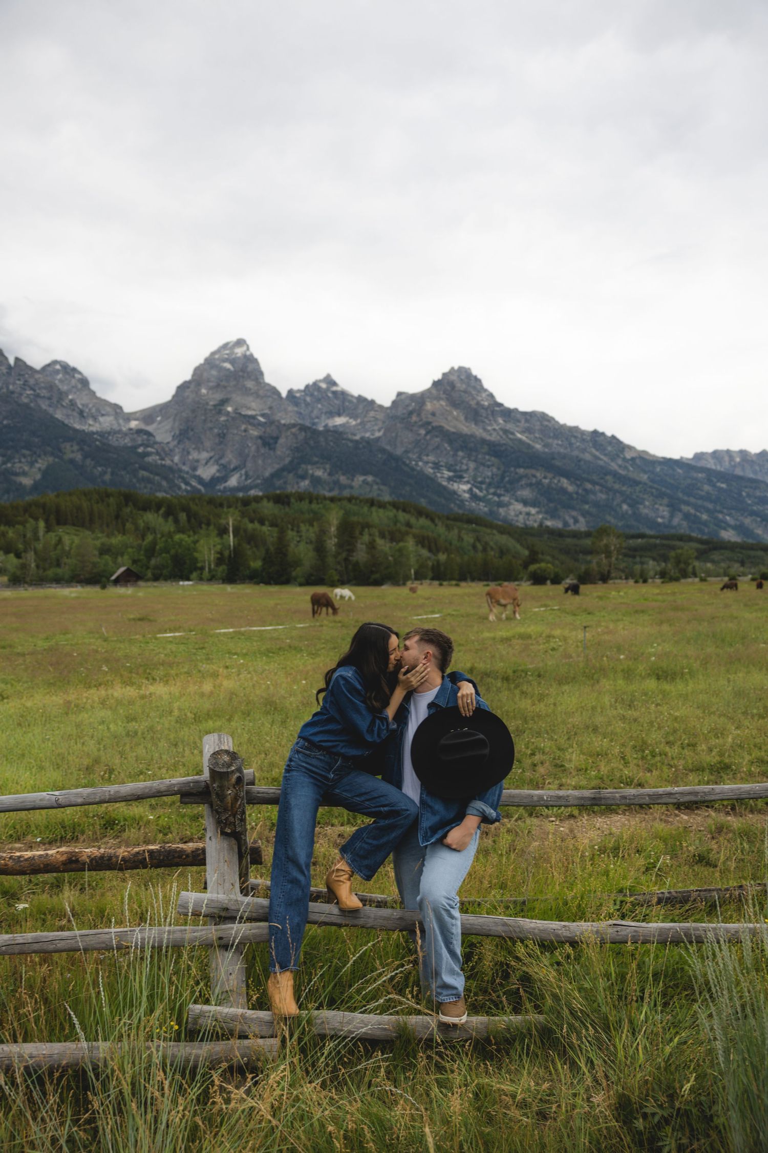 Couple sharing a romantic kiss on a rustic fence with Grand Teton mountains and green meadow in background.