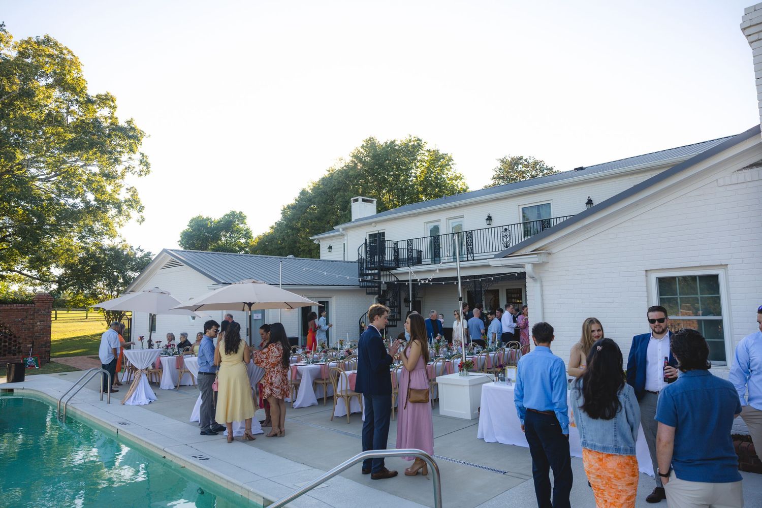 Elegant outdoor garden party with guests mingling poolside at a white estate home during a summer evening event.