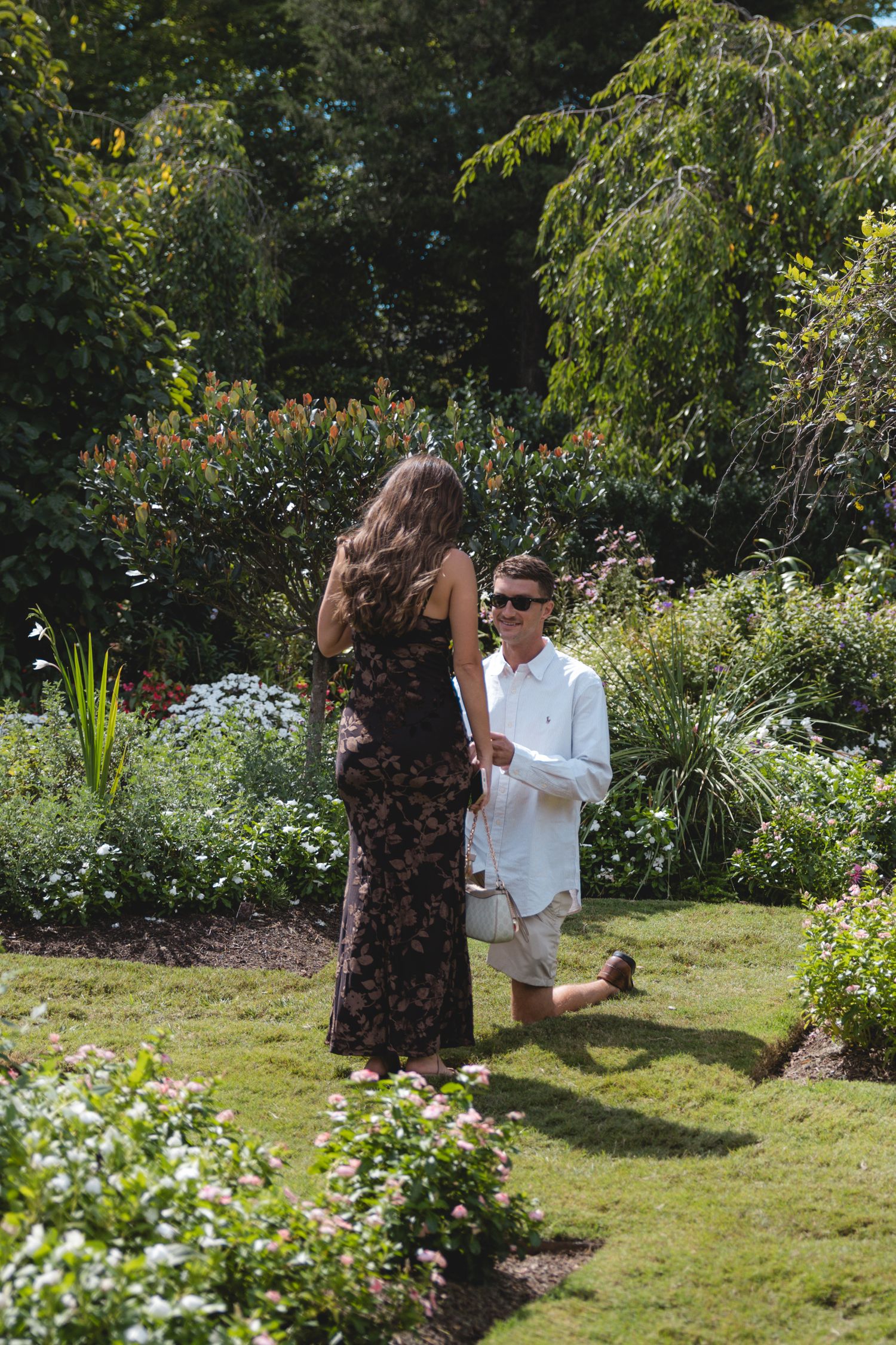 Man kneeling on one knee proposing to woman in floral dress in a lush garden on a sunny day.