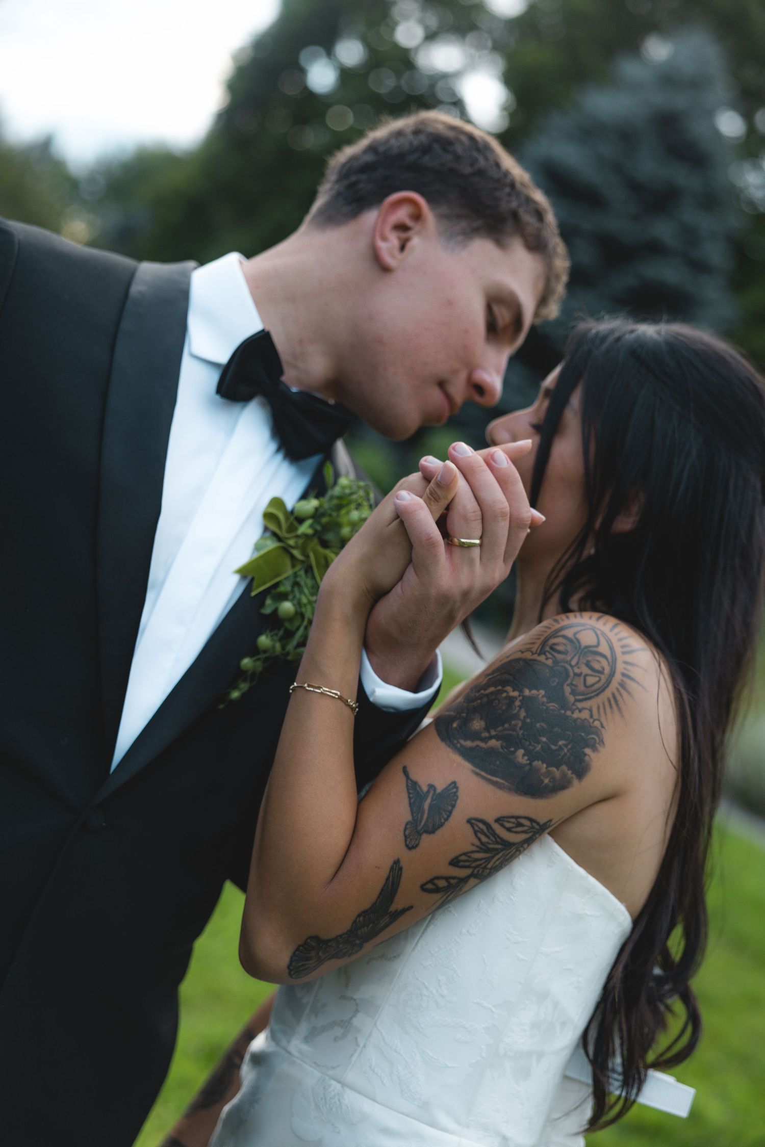 A bride and groom share a romantic moment, holding hands and nearly kissing, she wearing a white gown with tattoos.