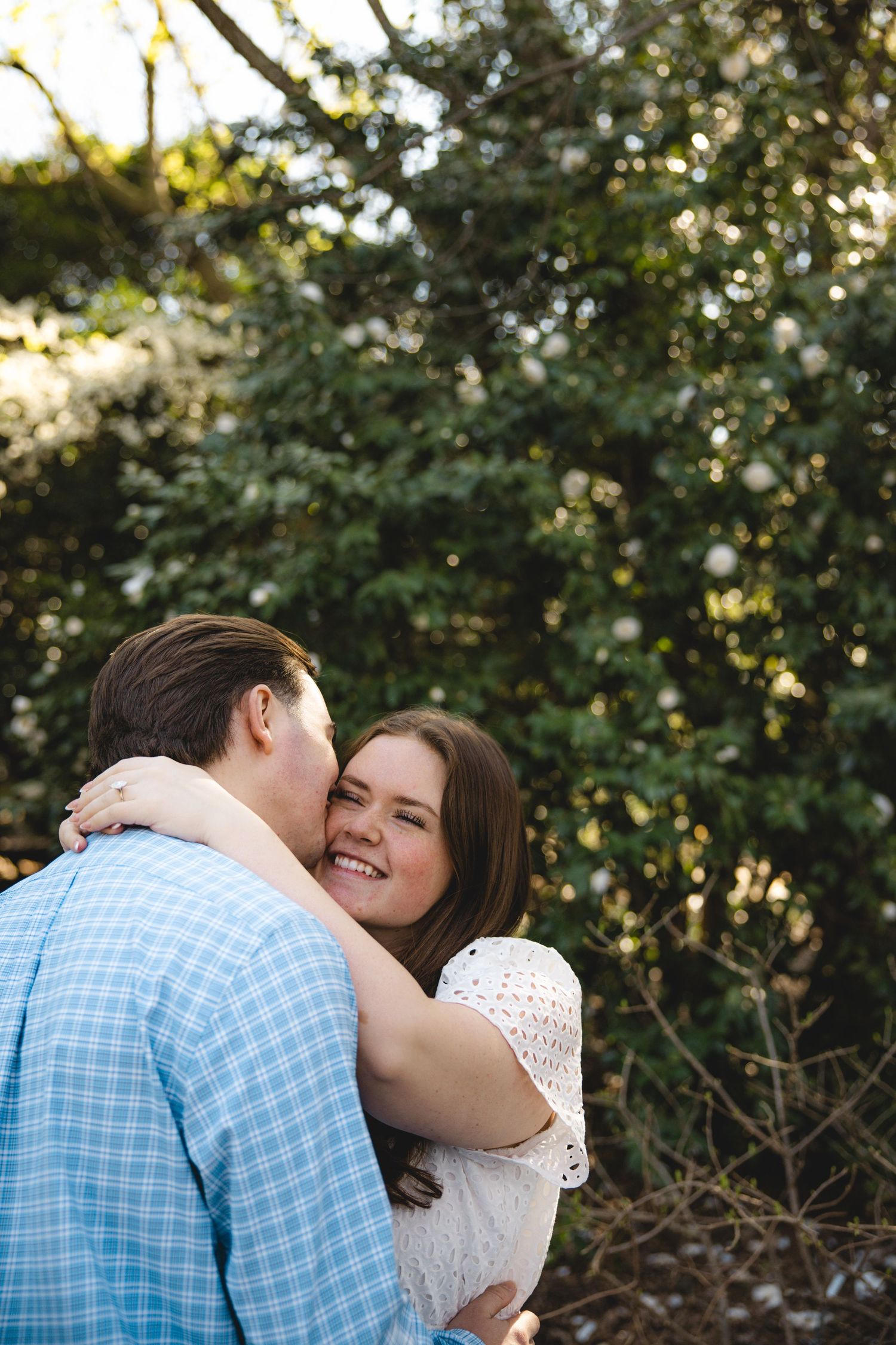 A couple embraces outdoors near blooming white flowers, the woman smiling as her partner kisses her cheek.