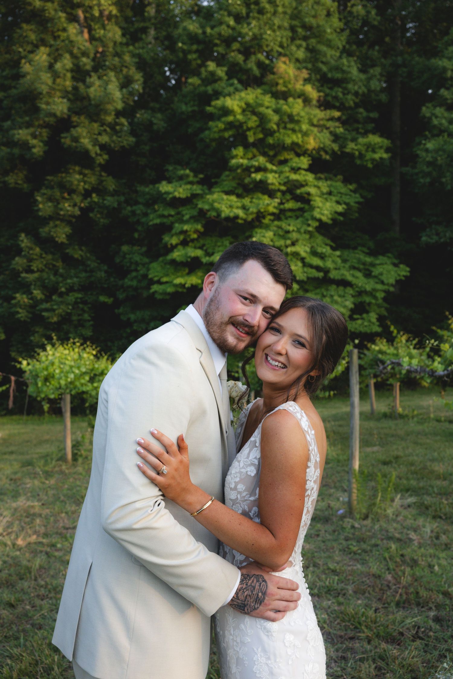 A smiling bride and groom embrace outdoors in a lush green vineyard setting, she in lace gown, he in cream suit.