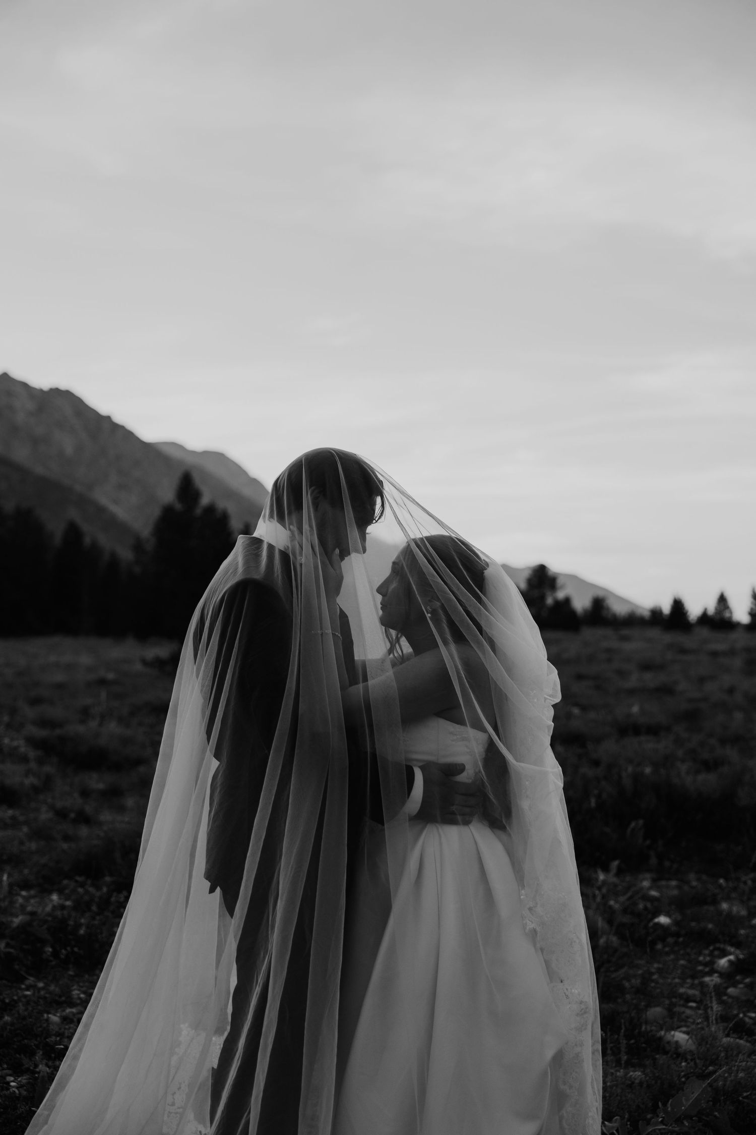 Black and white photo of a bride and groom sharing a kiss under a flowing veil with mountain scenery behind them.