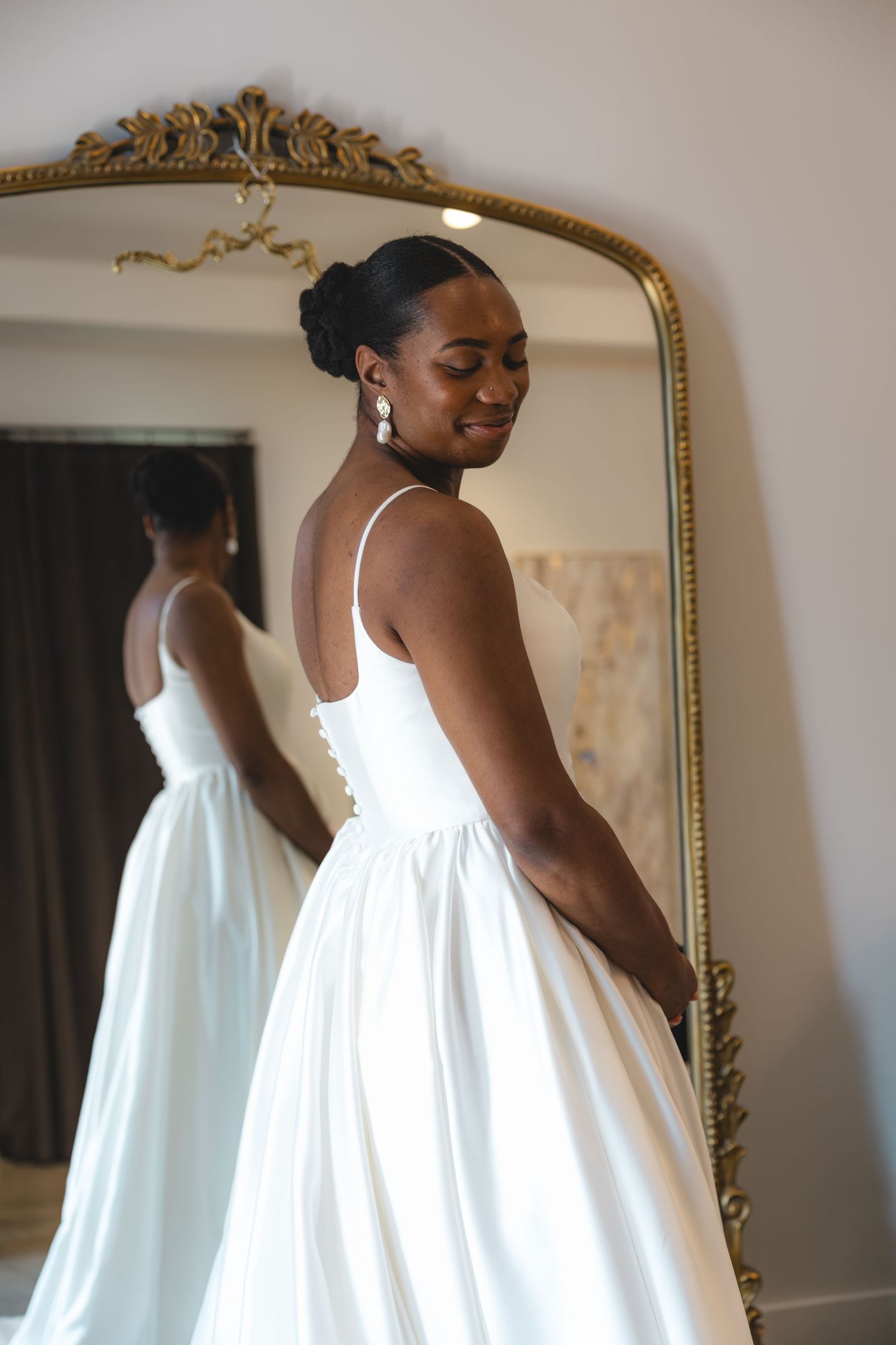 Bride in elegant white satin ball gown with spaghetti straps, smiling beside ornate gold mirror in bridal boutique.