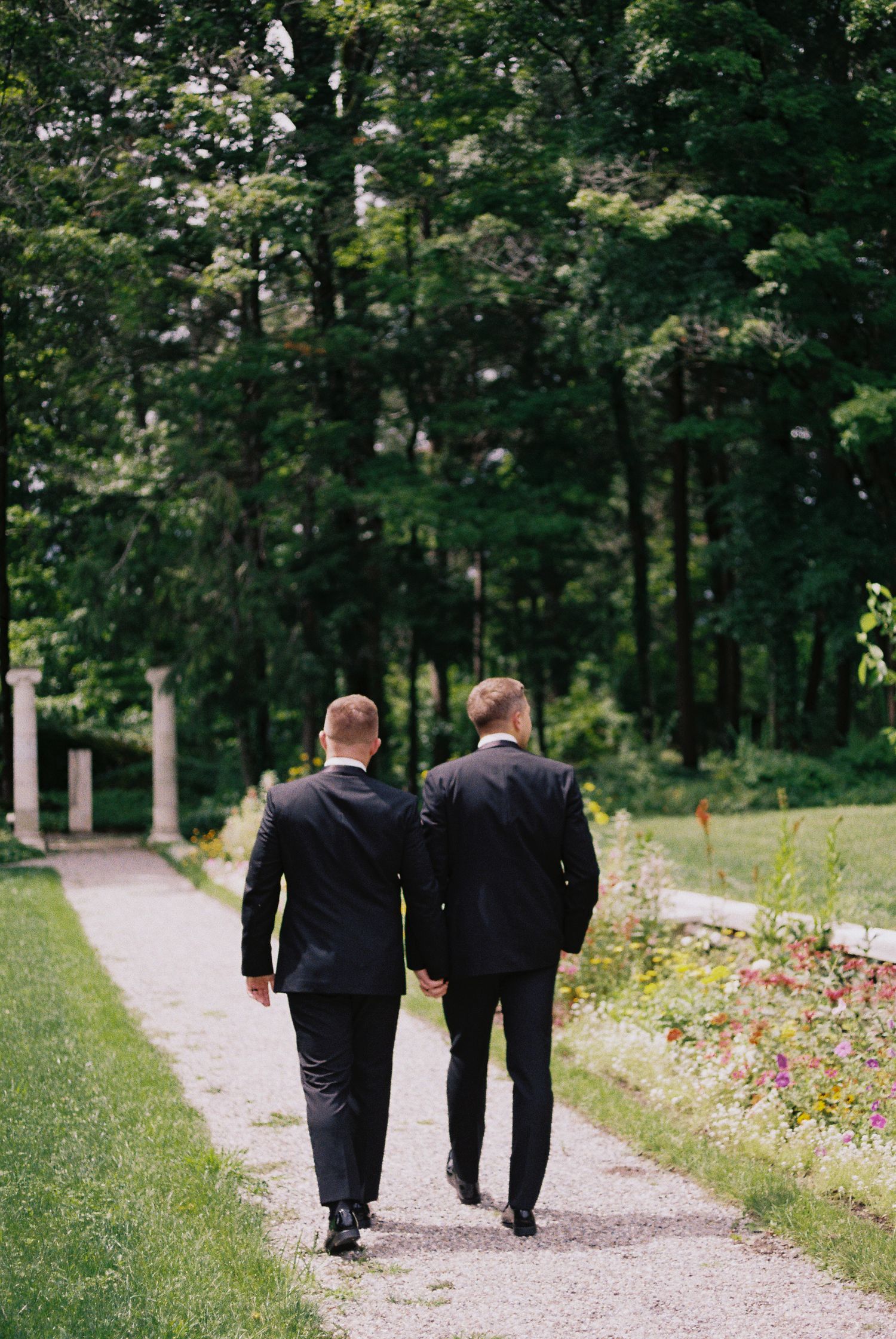 Two grooms in black suits walk hand in hand down a garden path surrounded by lush green trees.